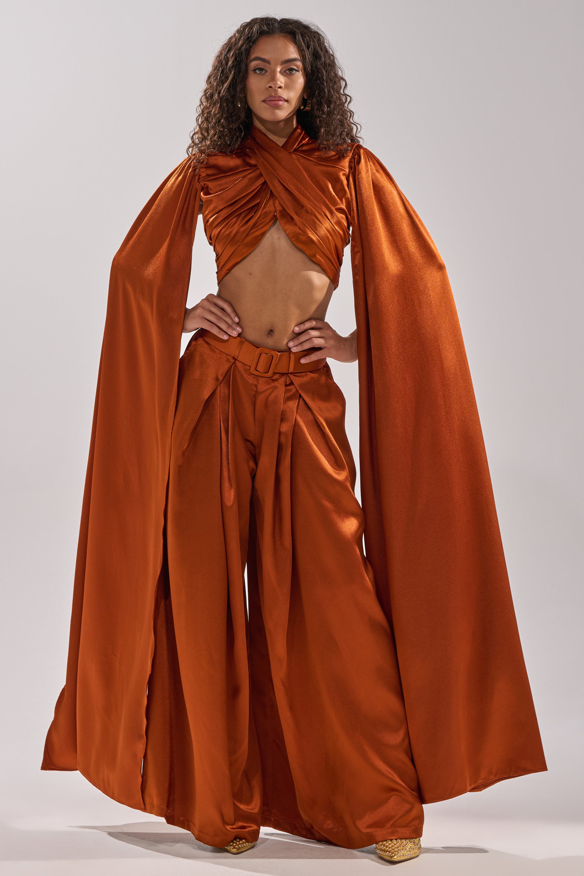 A woman poses confidently in the ALL WE EVER KNEW BLOUSE by AKIRA Label, featuring rust-orange satin, a cropped fit, wide-leg pants, and dramatic floor-length cape sleeves. Her curly hair is loose against a light background.