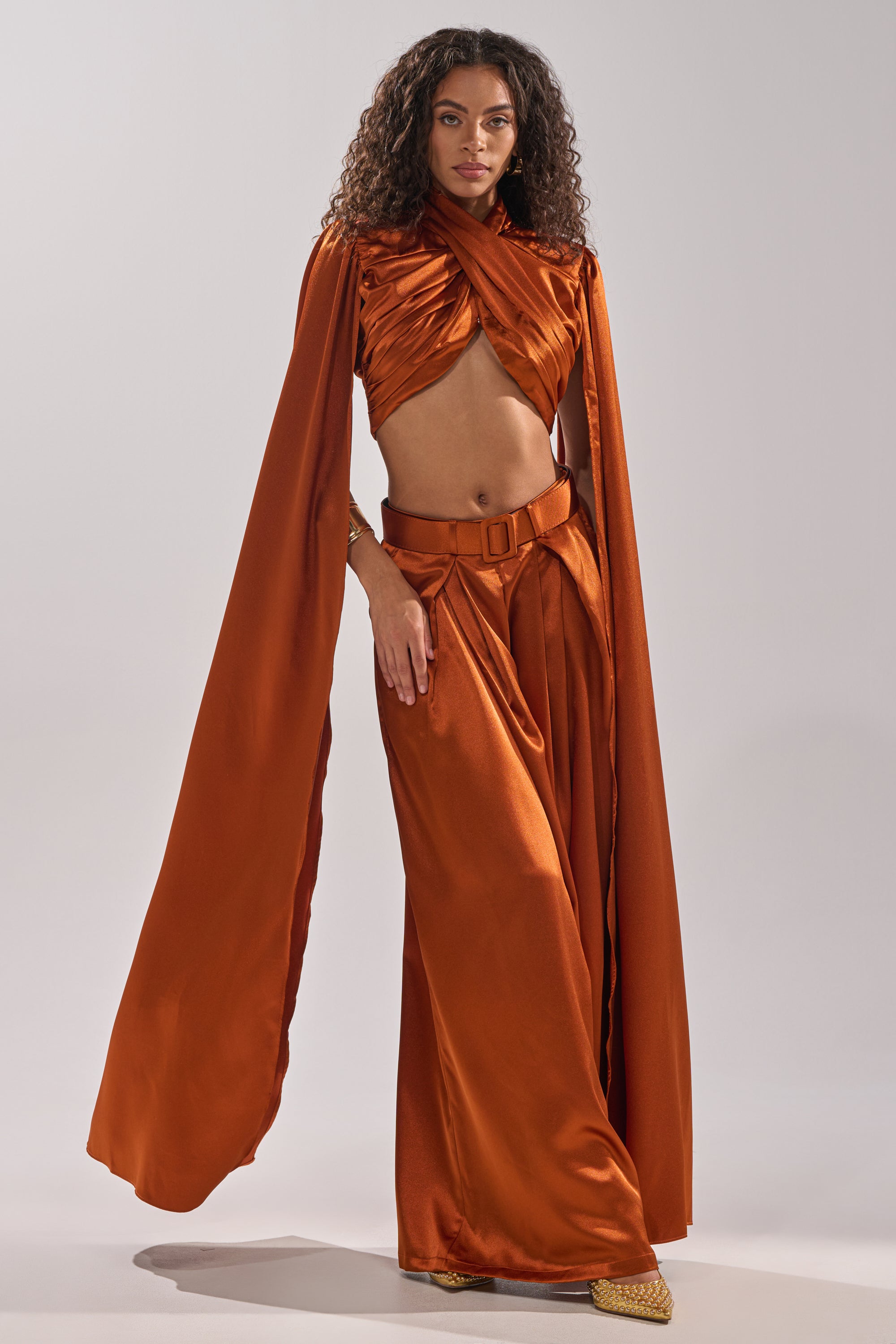 A woman stands confidently against a plain background in the ALL WE EVER KNEW BLOUSE, a shiny burnt orange AKIRA Label top with a cropped fit, long flowing cape sleeves, paired with high-waisted wide-leg pants and yellow pointed shoes.