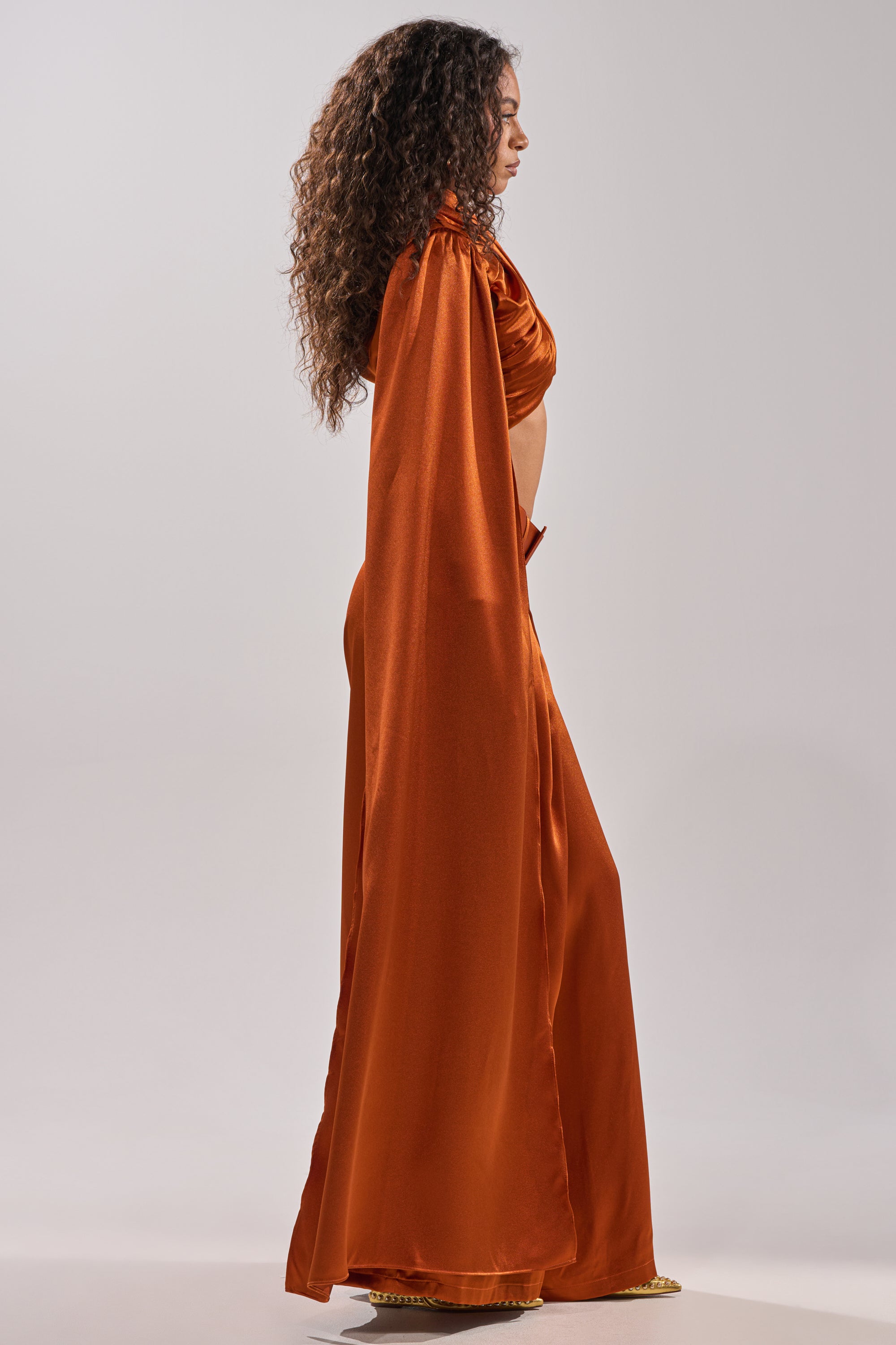A woman with long curly hair stands in profile, wearing the ALL WE EVER KNEW BLOUSE by AKIRA Label—a flowing burnt orange top with a cape-like sleeve and cropped fit—set against a plain, light-colored background.