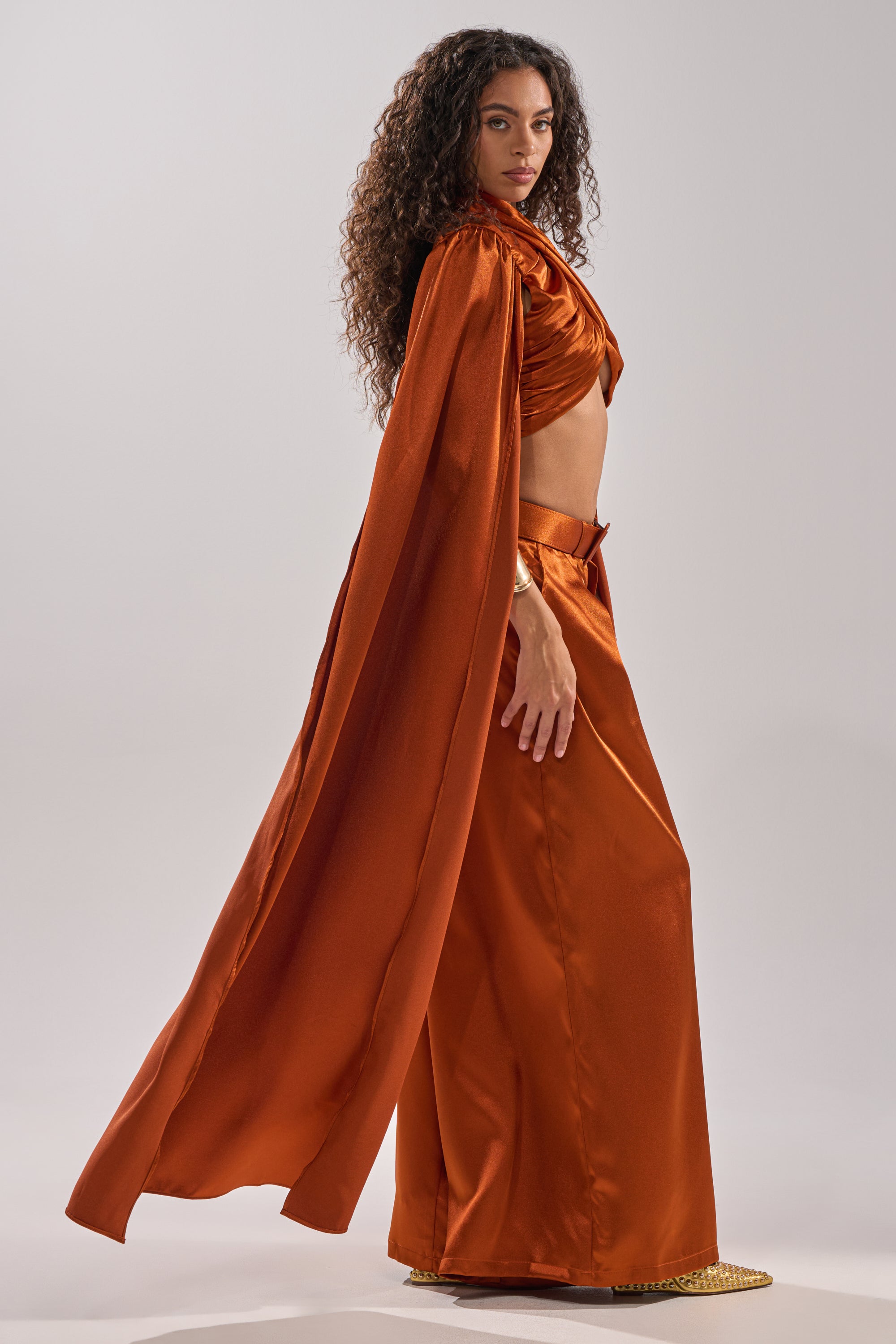 A woman with long curly hair wears the shiny, burnt orange ALL WE EVER KNEW BLOUSE by AKIRA Label, featuring a cropped fit and cape, paired with wide-leg pants as she stands in profile against a plain light background.