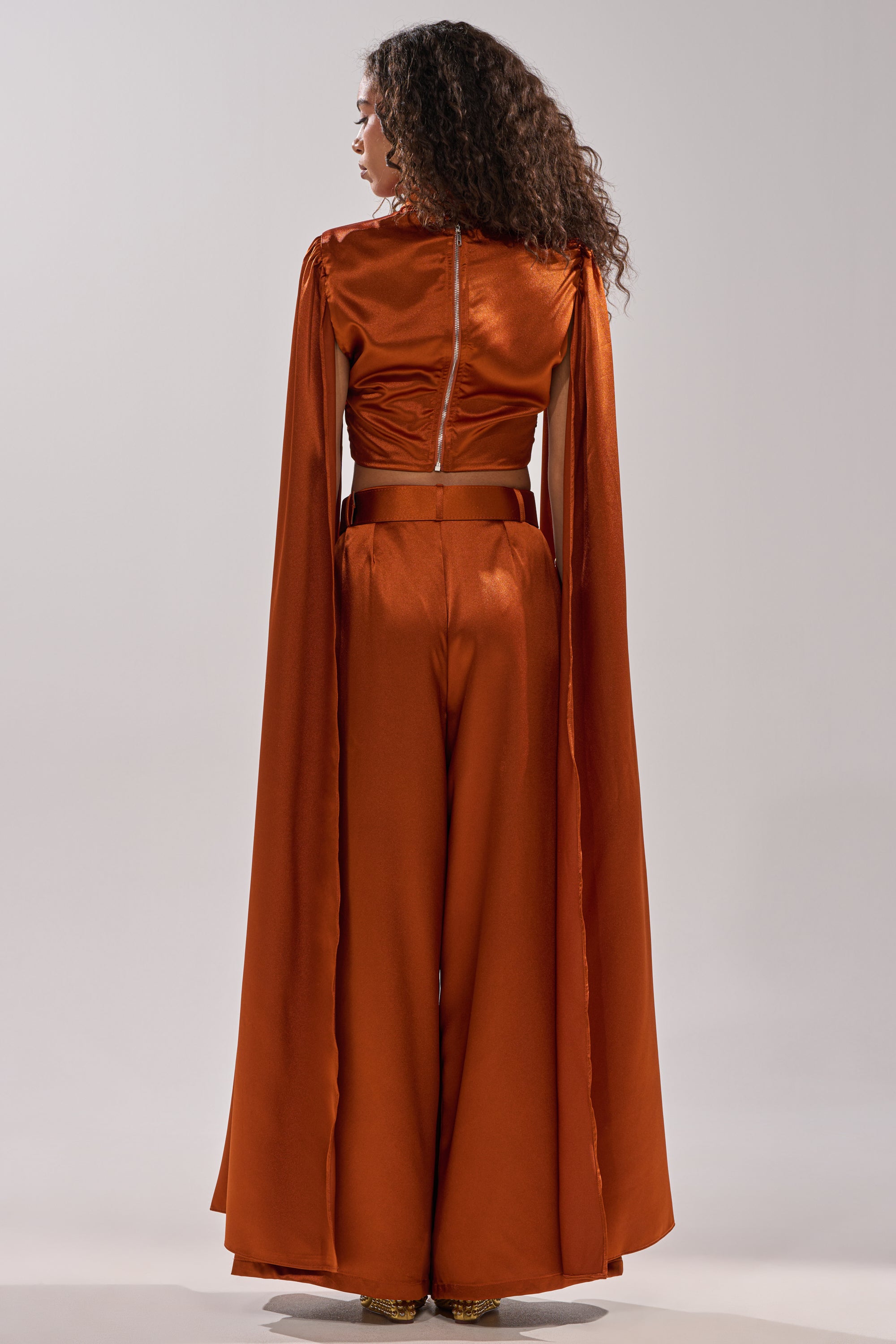 A woman with curly hair faces away, wearing the shiny burnt orange ALL WE EVER KNEW BLOUSE by AKIRA Label. The cropped blouse features wide-leg pants and long cape-like sleeves, set against a plain light background.