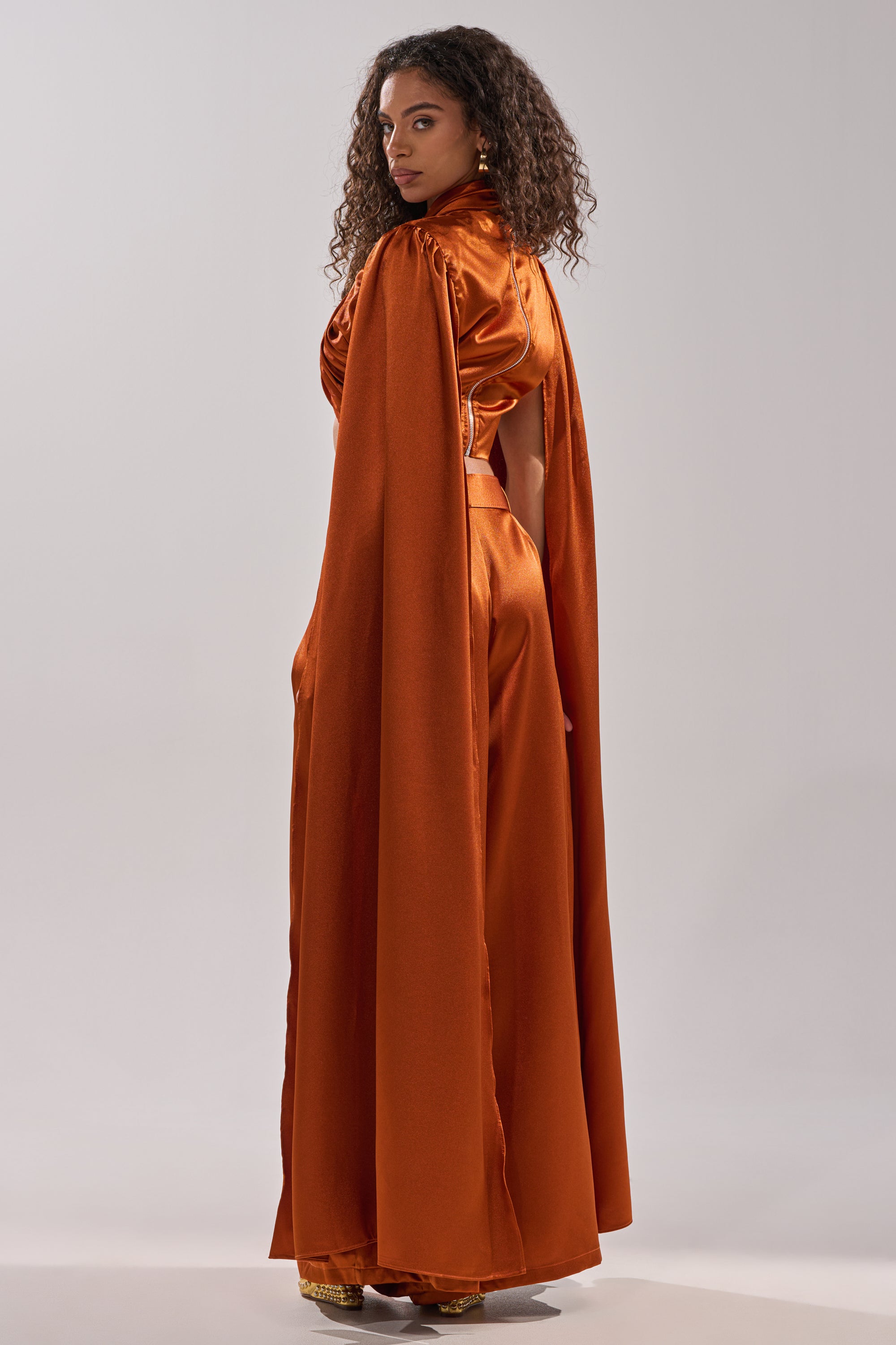 A woman with curly hair stands sideways against a plain light background, wearing the AKIRA Label ALL WE EVER KNEW BLOUSE in shiny orange satin, paired with wide-legged pants and a matching cape, looking over her shoulder.