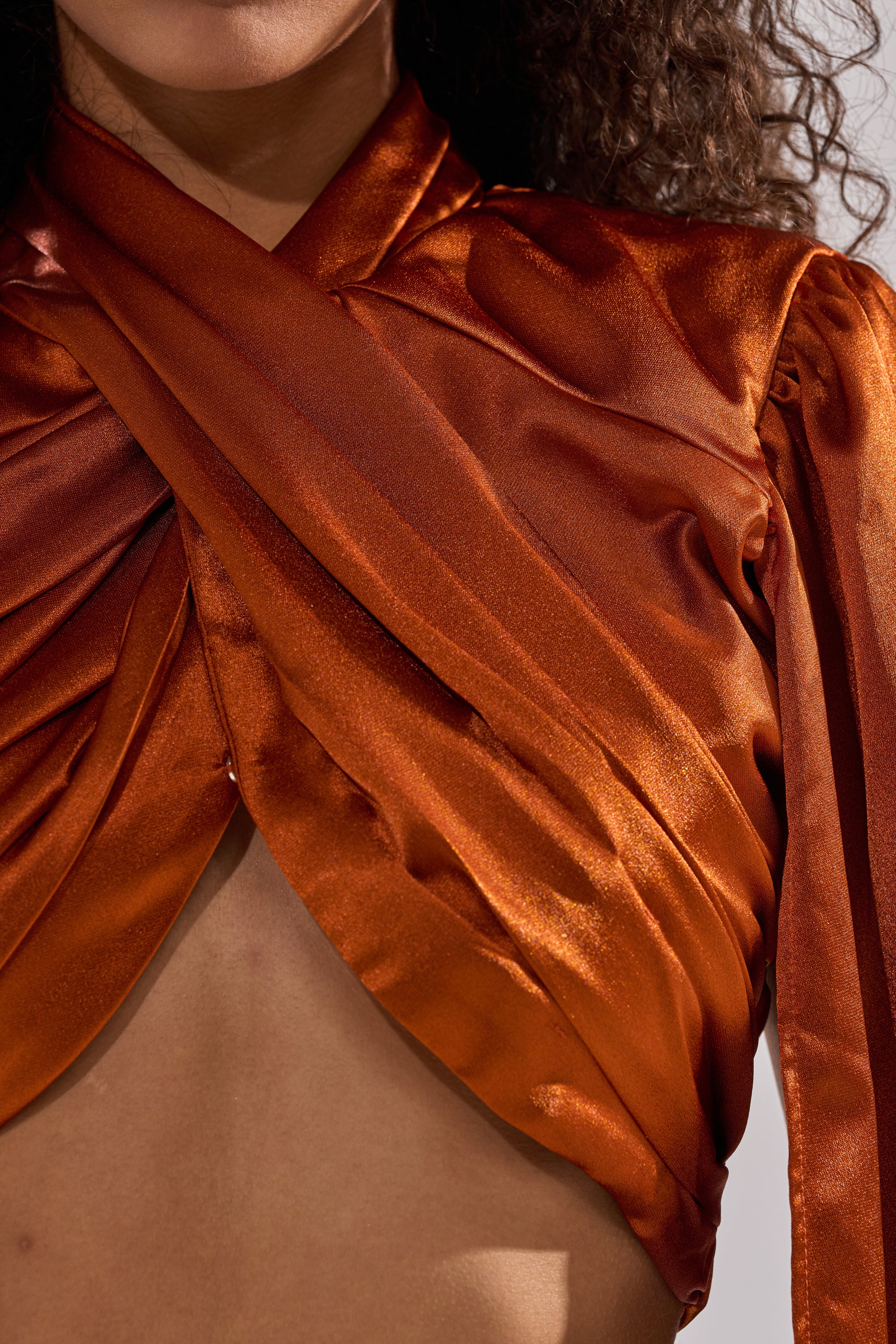 A close-up of a person wearing the shiny, rust-colored ALL WE EVER KNEW BLOUSE featuring a wrapped, crisscross design and cropped fit that reveals part of the chest. Only the lower face, neck, and some curly hair are visible.
