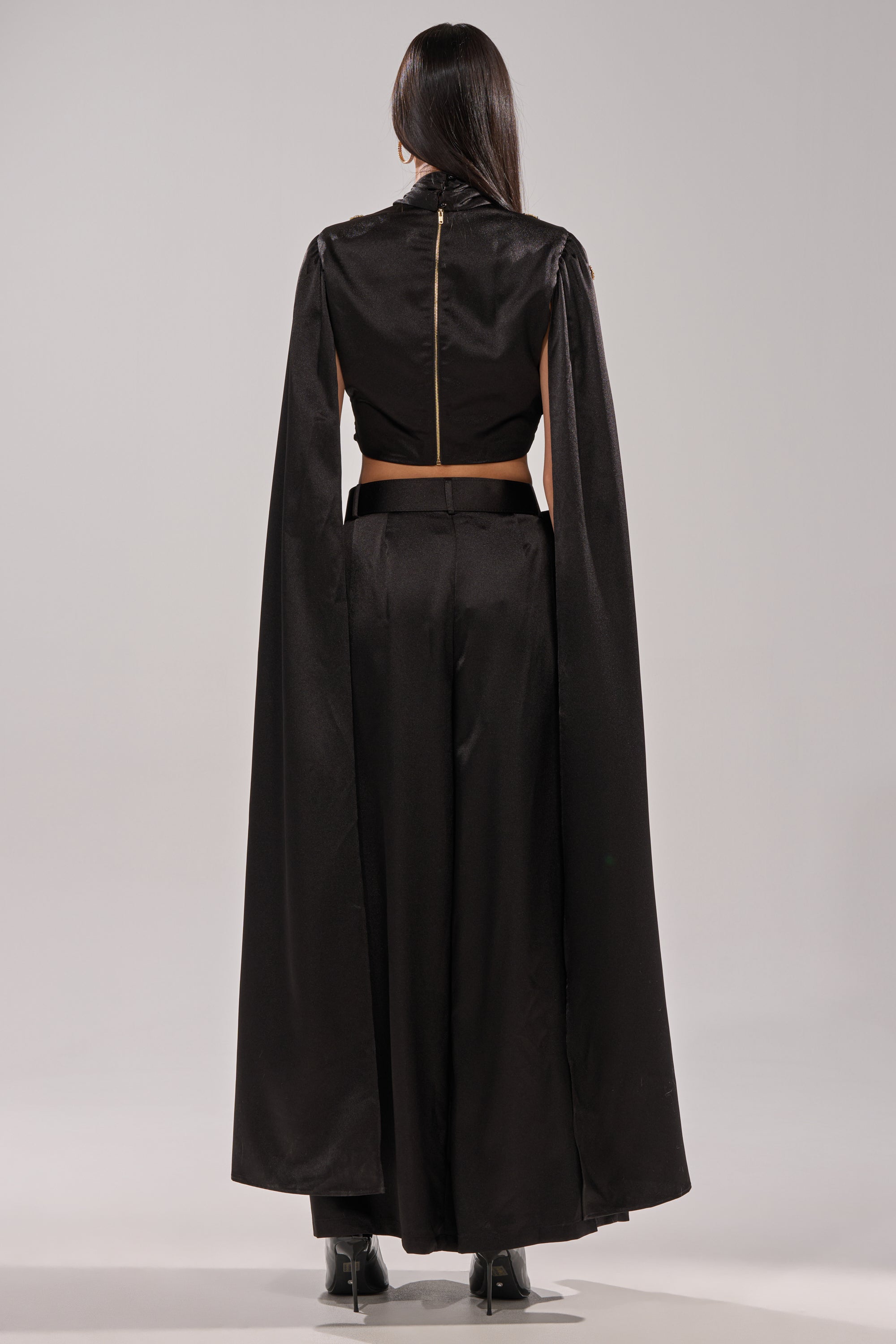 A person with long dark hair faces away, wearing the AFTER DARK BLOUSE—a black crop top with a gold zipper—paired with high-waisted black pants and a long cape adorned with gemstone appliqués. The backdrop is plain and light-colored.