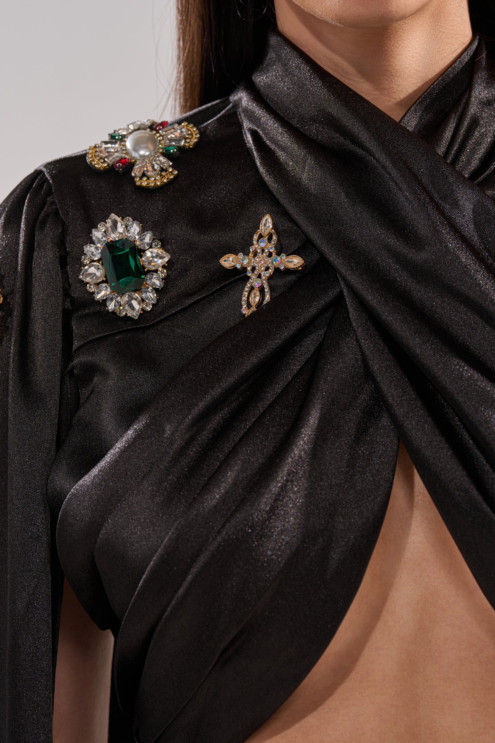 A close-up of a woman wearing the AFTER DARK BLOUSE, a shiny black satin top featuring ornate jeweled brooches, green and pearl gemstone appliqués, and a gold cross-shaped brooch, with part of her chest visible.