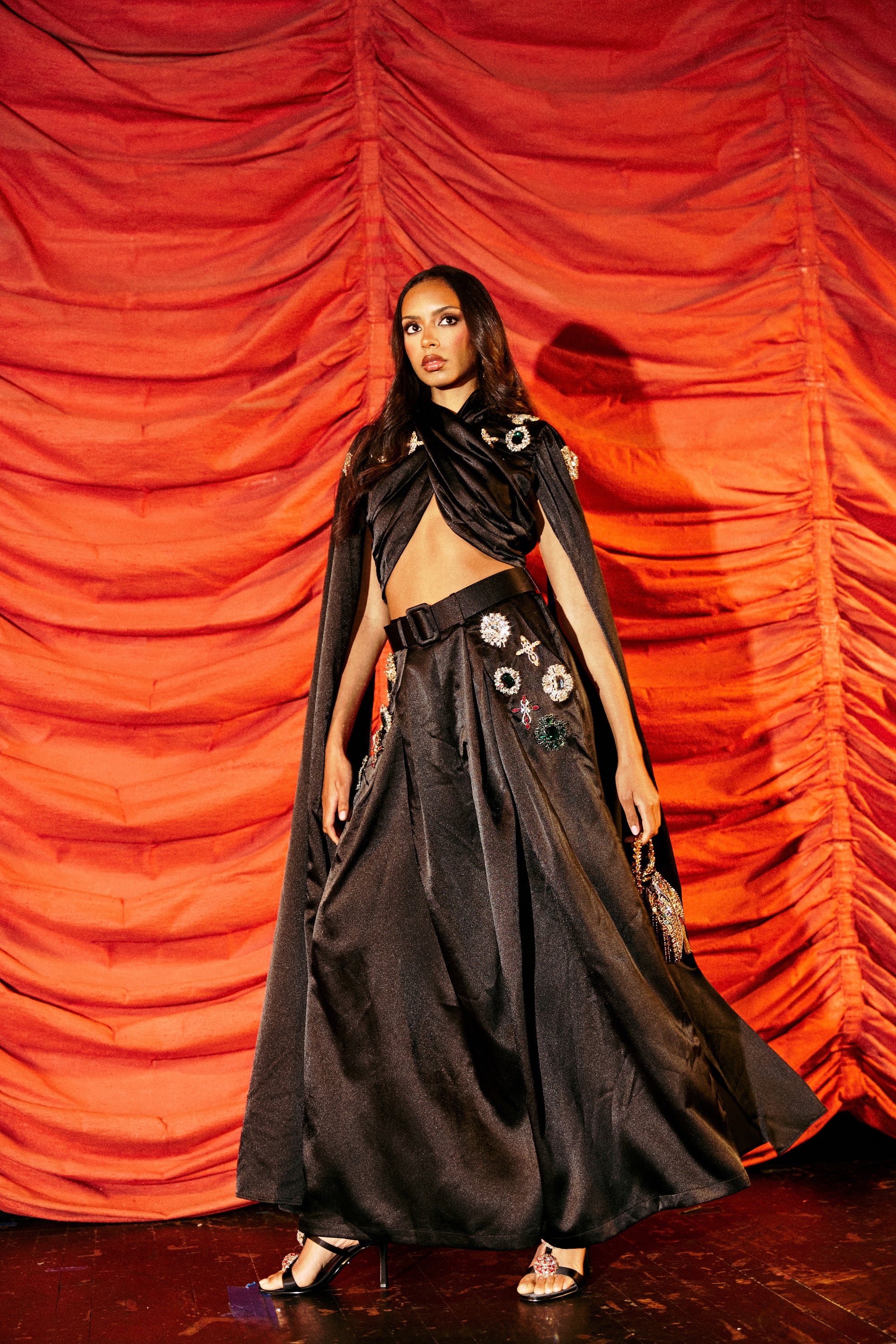 A woman poses before red curtains wearing a black floor-length skirt, an AFTER DARK BLOUSE with gemstone embellishments, a black cape, and black high-heeled sandals.