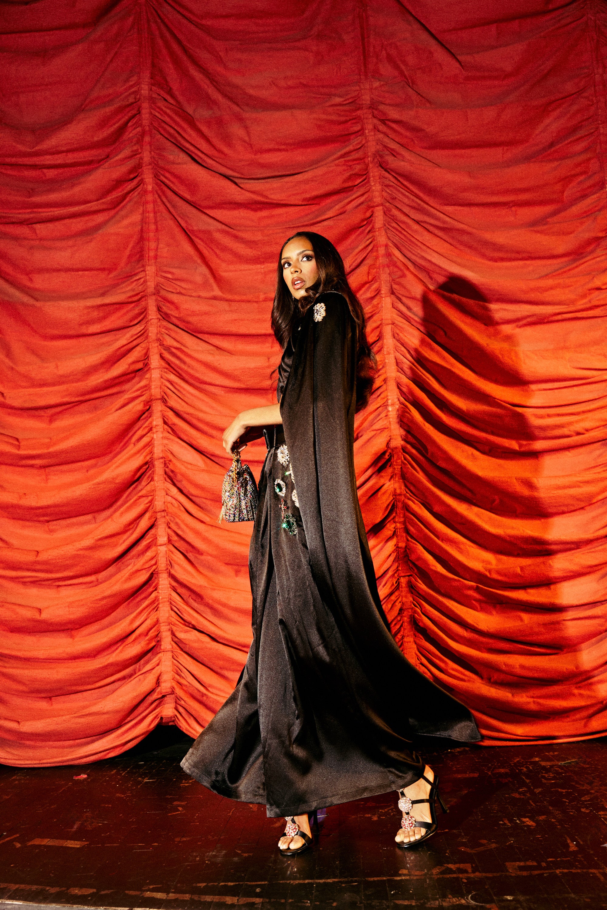 A woman wearing the AFTER DARK BLOUSE, a gemstone-embellished top, pairs it with a flowing black gown and high heels as she walks confidently onstage in front of red curtains, carrying a small handbag and glancing to the side.