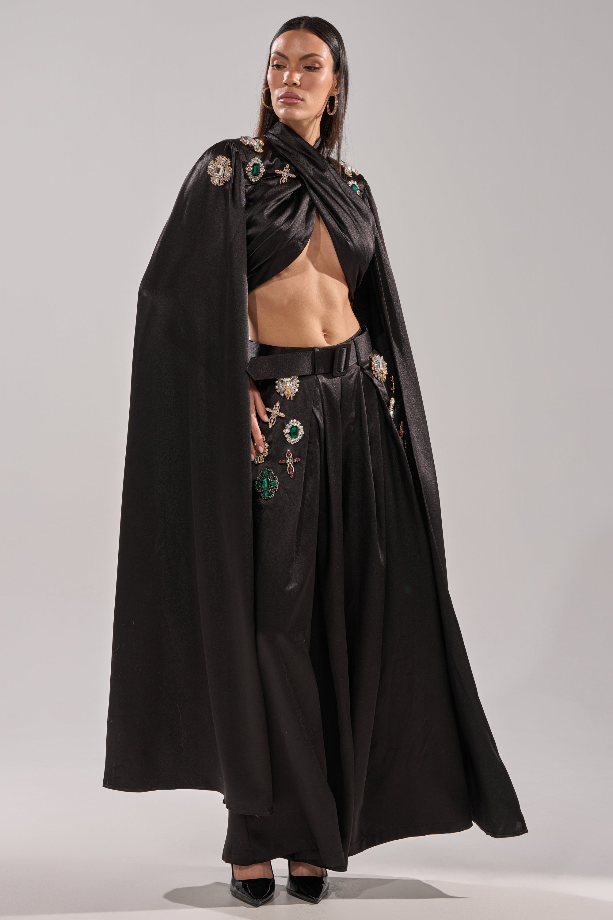 A person stands confidently in the AFTER DARK BLOUSE, styled with a black, floor-length floral-embellished cape and wide-leg pants. Their long hair is center-parted, and they wear black shoes against a plain, light-colored background.