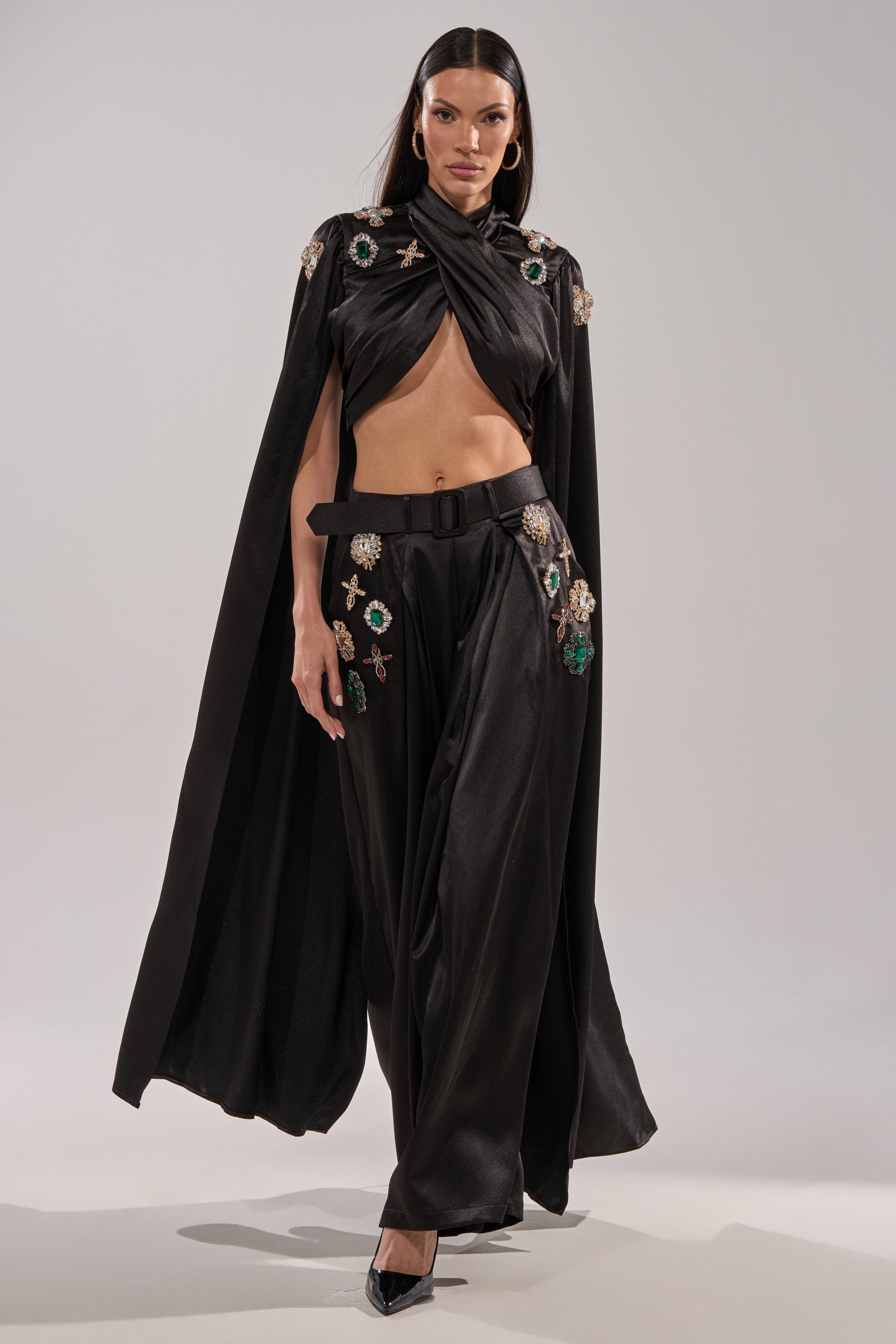 A woman poses confidently in black heels, wearing the AFTER DARK BLOUSE—a black cape-style top with a twisted front that exposes her midriff—paired with wide-legged black pants featuring vibrant gemstone appliqués.