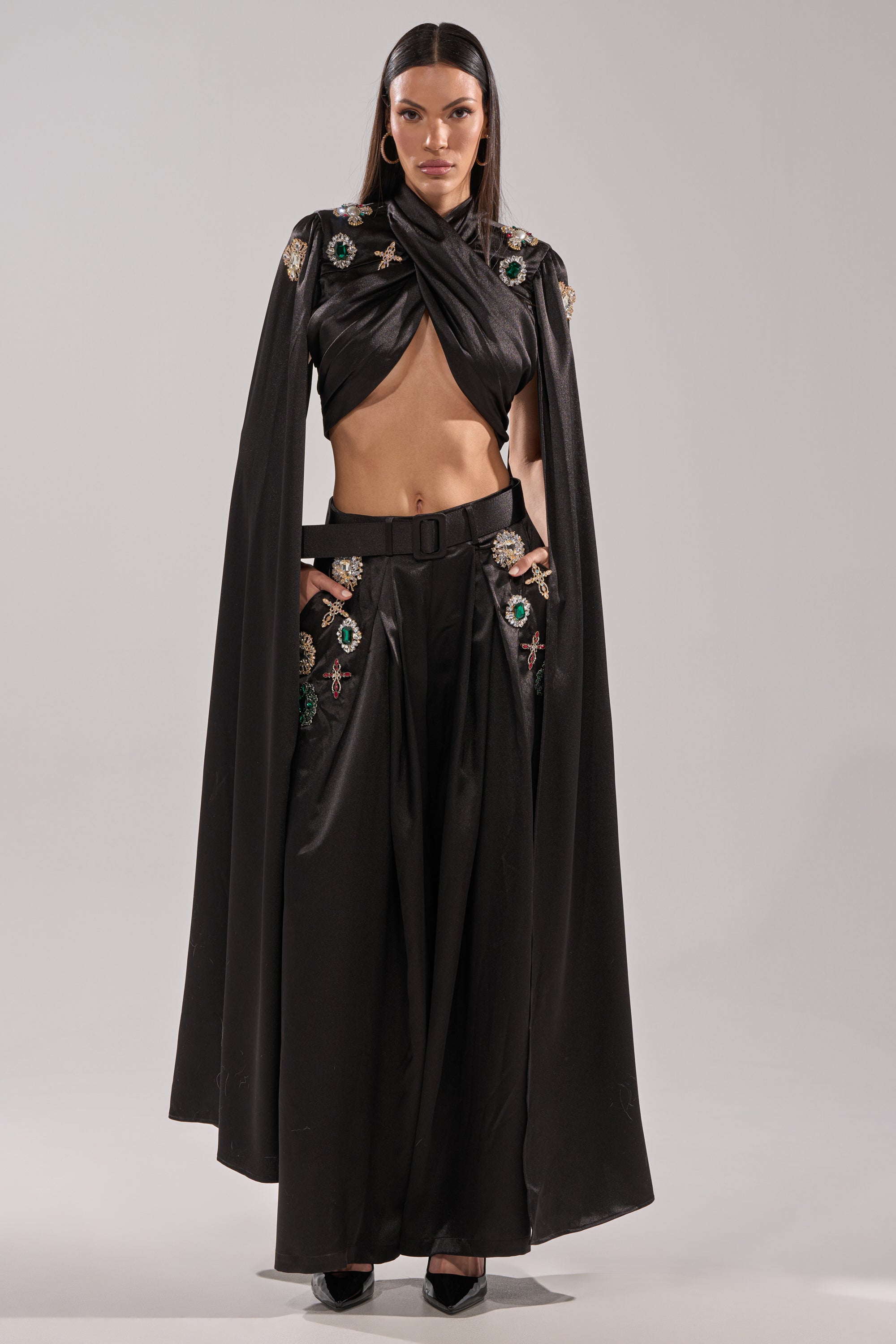 A woman poses facing forward in the AFTER DARK BLOUSE and wide-legged pants with colorful embroidered patches, accessorized with hoop earrings and black heels. She has long straight hair and stands against a plain light background.