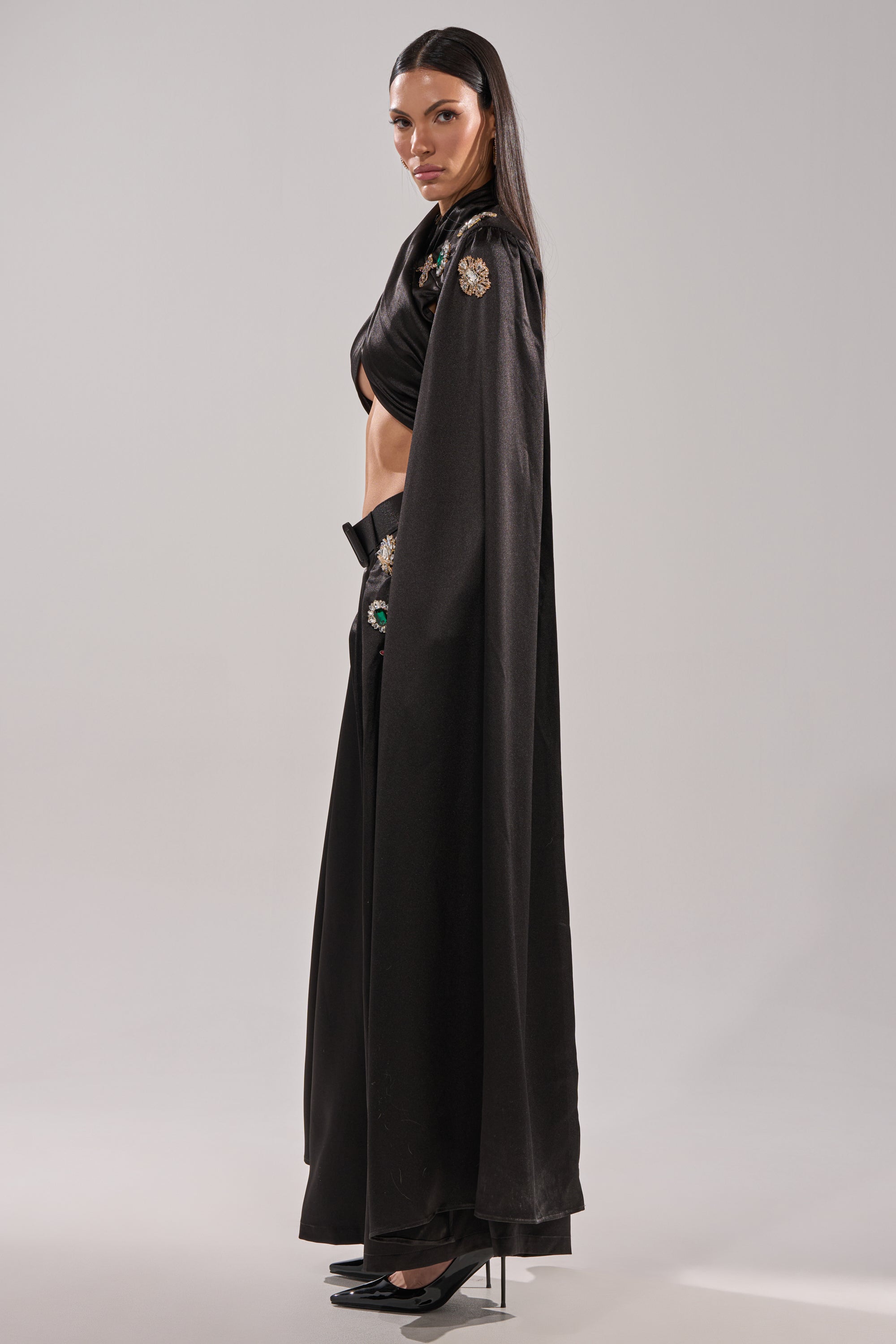 A woman stands in profile against a plain light background, wearing the AFTER DARK BLOUSE — a cropped black top paired with black pants, high heels, and a long black cape adorned with gemstone appliqués.