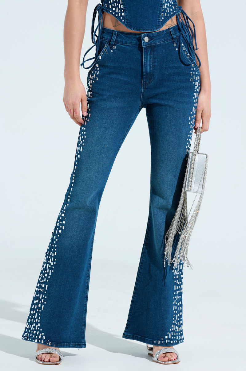 Rocking Rockstar Denim, someone showcases high-waisted, flared blue jeans with rhinestone accents. They wear a matching top with string details at the waist and carry a silver fringed purse against a plain light background.