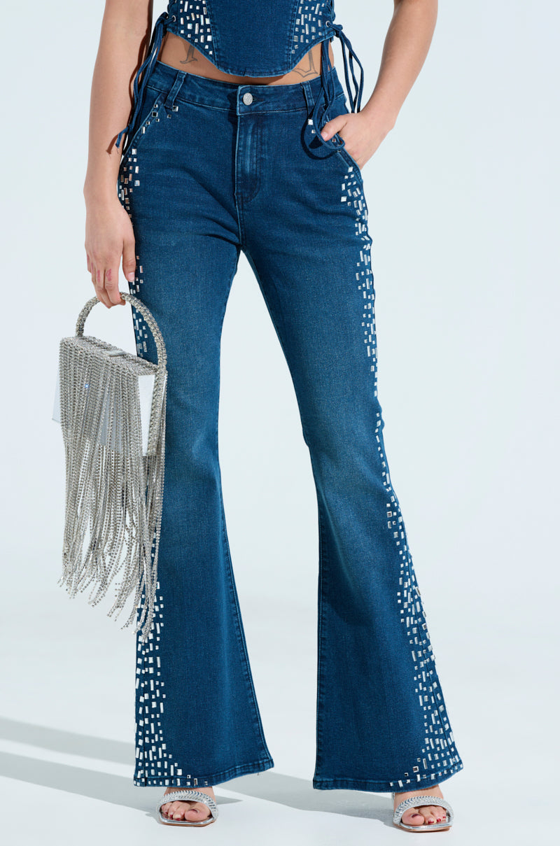 A fashionista wears Rockstar Denim Rhinestone Flared Pants with white square details, paired with a chic top. They accessorize with a silver fringed handbag and open-toed shoes against a light, neutral background.