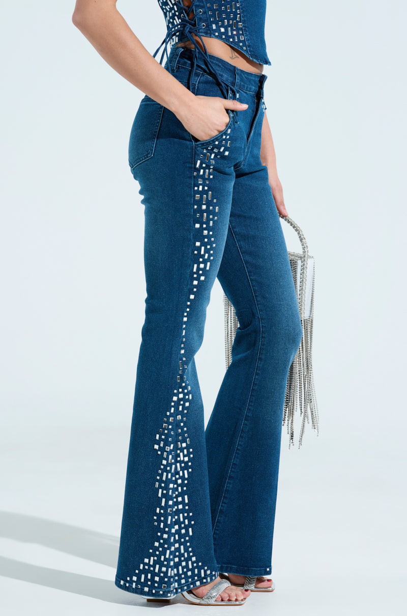 Someone wearing ROCKSTAR DENIM RHINESTONE FLARED PANT with a sparkling geometric pattern on the side seam holds a silver fringe handbag and wears silver sandals.