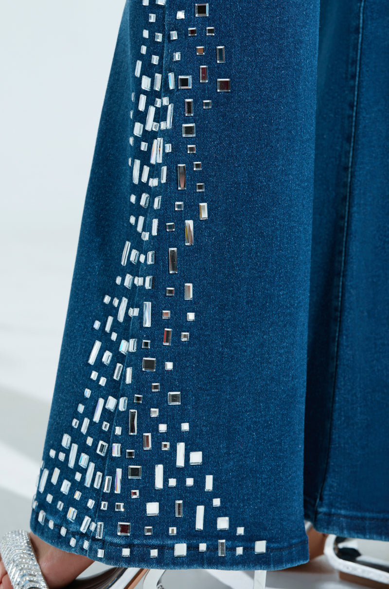 A close-up of the ROCKSTAR DENIM RHINESTONE FLARED PANT shows a dark blue fabric adorned with reflective square and rectangular embellishments in a scattered pattern, complemented by a visible silver high-heeled shoe, echoing AKIRA Label’s signature style.