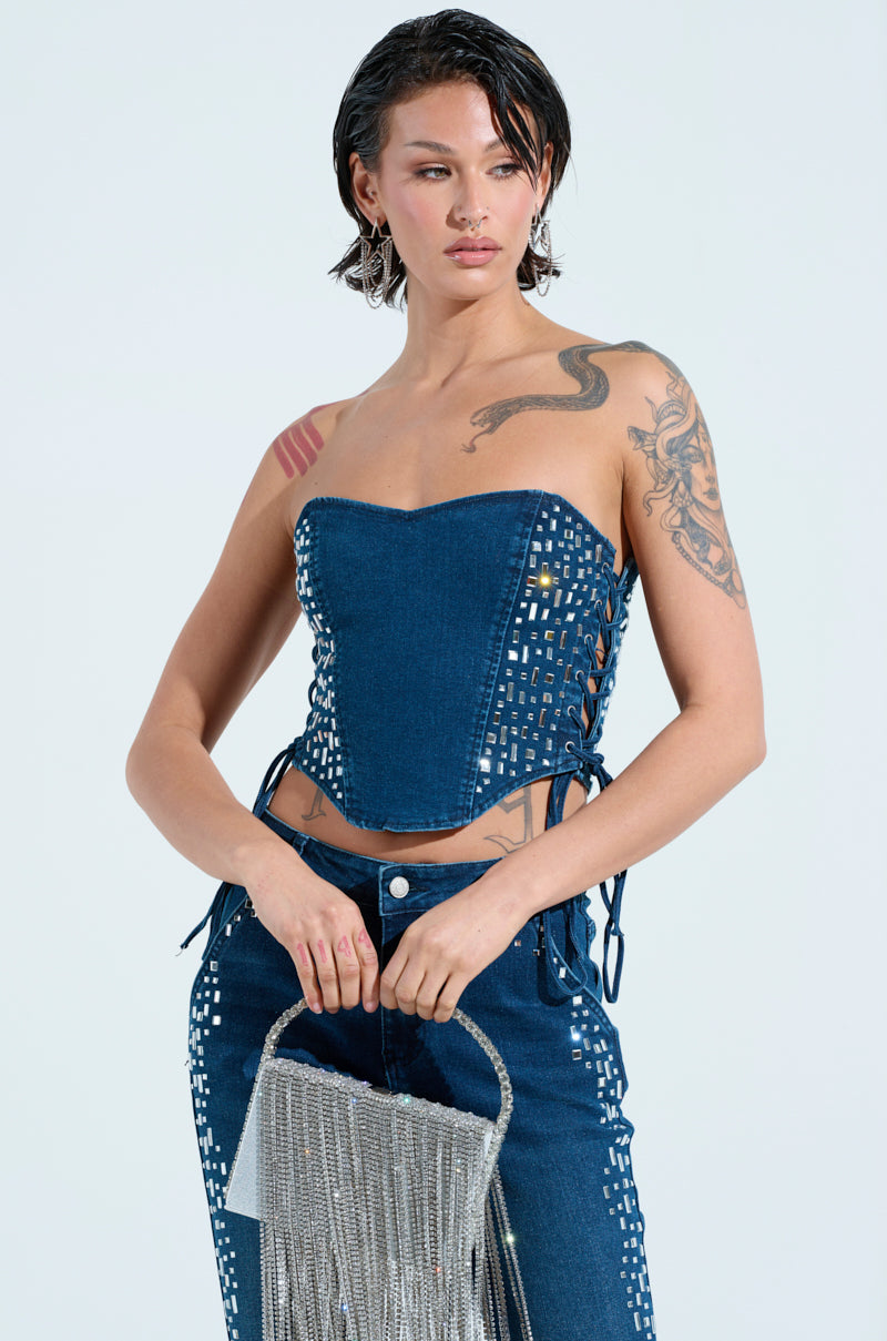 A person flaunts the ROCKSTAR DENIM RHINESTONE CORSET TOP paired with matching pants, holding a fringed silver handbag. With tattoos and short, wet-look hair against a light background, they effortlessly exude VIP access glamour.