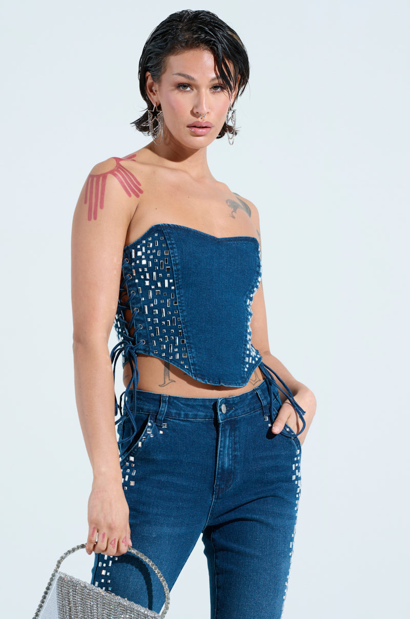 A person with short hair wears the ROCKSTAR DENIM RHINESTONE CORSET TOP and jeans, beautifully adorned with silver studs. They're holding a metallic handbag and have a chest and shoulder tattoo against a light blue background, evoking an exclusive VIP access vibe.