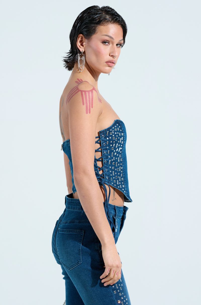 With short dark hair, this person sports a ROCKSTAR DENIM RHINESTONE CORSET TOP featuring lace-up sides, matching denim jeans, and large hoop earrings. A red paint-like design decorates their shoulder against a soft light blue backdrop, enhancing their VIP vibe.