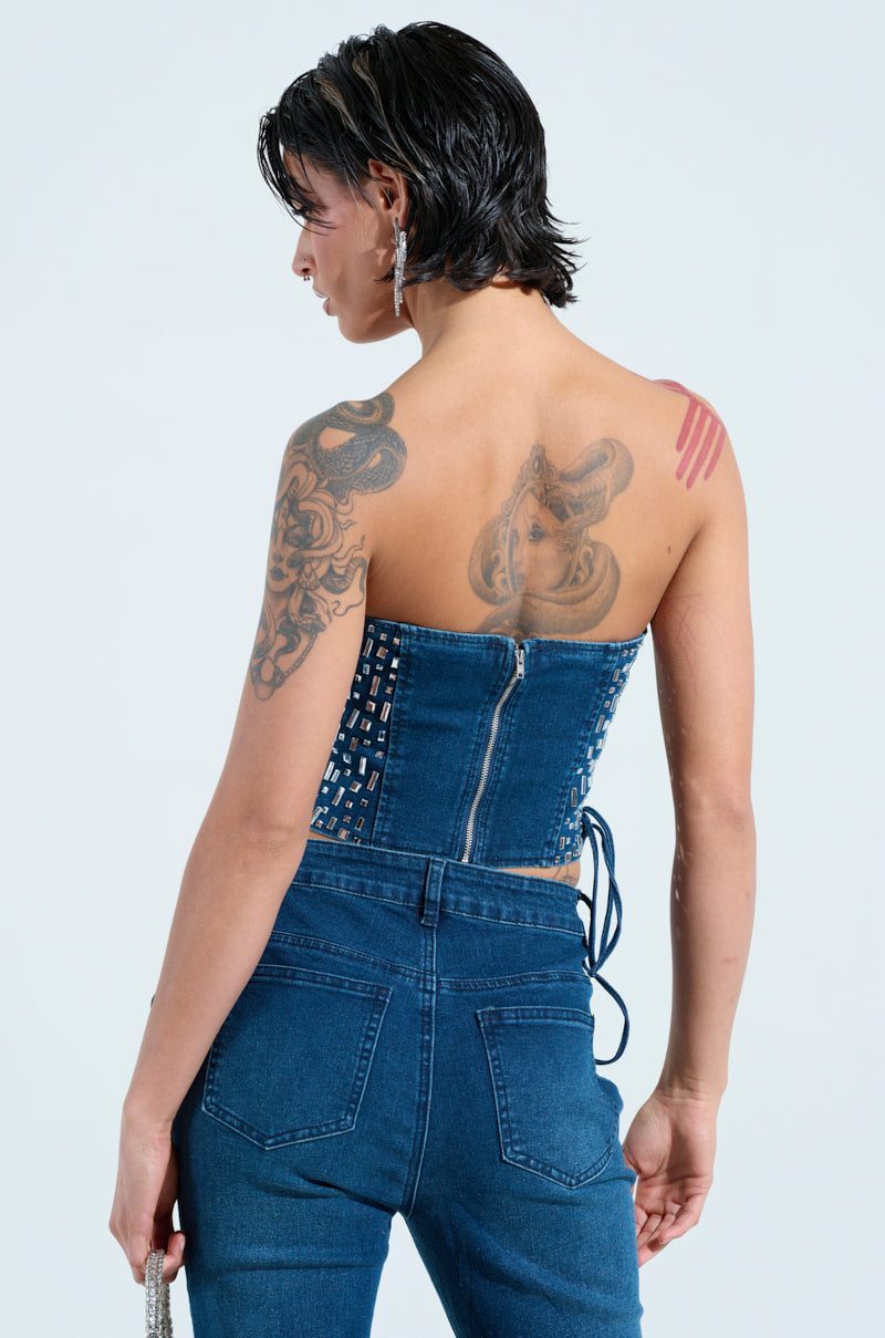 A person with short hair displays their tattoos while wearing the ROCKSTAR DENIM RHINESTONE CORSET TOP and jeans, slightly turned to the side. They have earrings, hold a small item in hand, and stand against a light background.