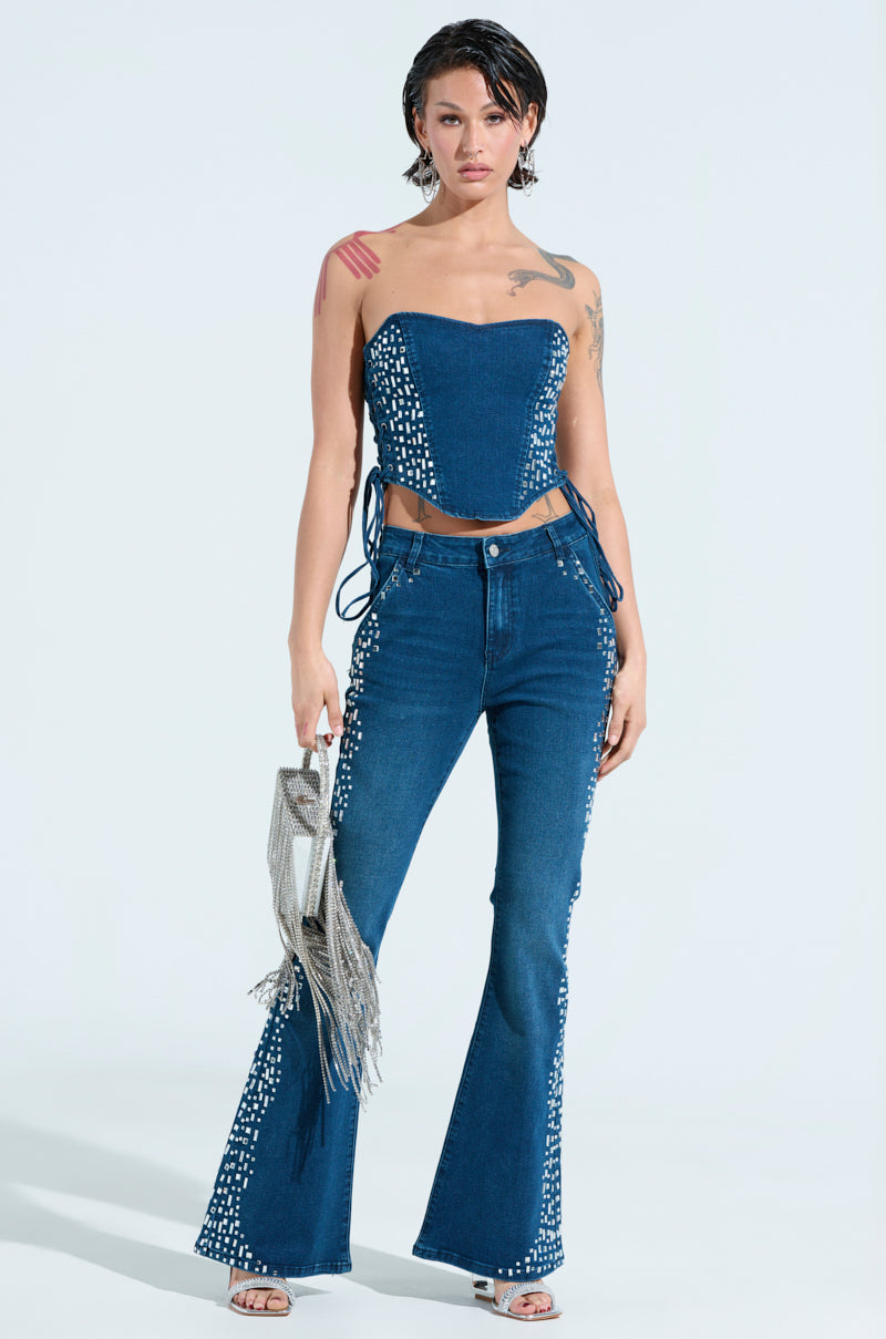 A person with short hair showcases their edgy style in a ROCKSTAR DENIM RHINESTONE CORSET TOP and flared jeans with white details, holding a silver fringed purse. Their tattoos stand out against the plain white background, enhancing their bold fashion statement.