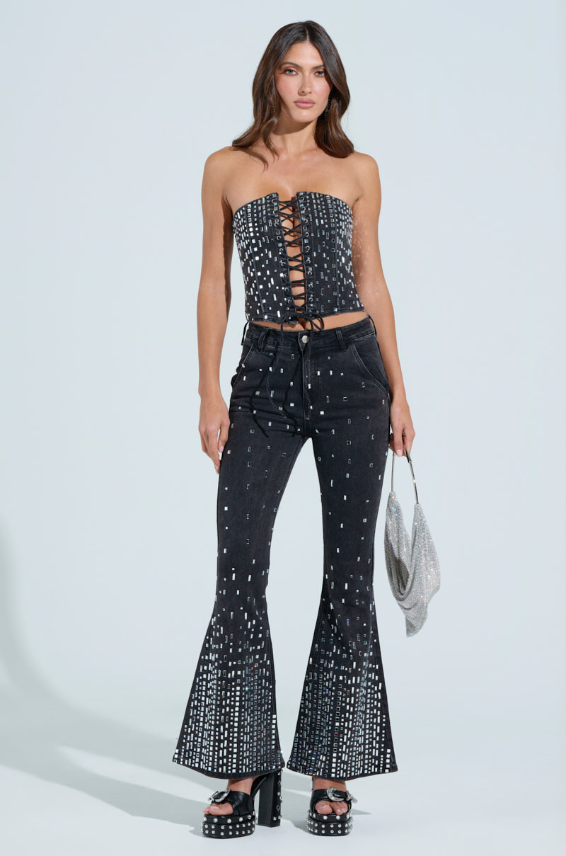 A woman wears a black strapless corset with lace-up front and POPSTAR BLACK DENIM PANT WITH RHINESTONE, both featuring white dot patterns. She accessorizes with a small silver bag, black platform heels, and her hair is styled in loose waves.