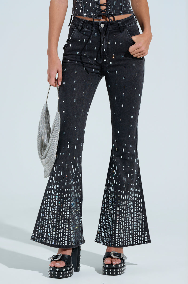 Someone dressed in POPSTAR Black Denim Pants with rhinestones, paired with a black lace-up top, holding a small silver handbag and wearing studded platform sandals.