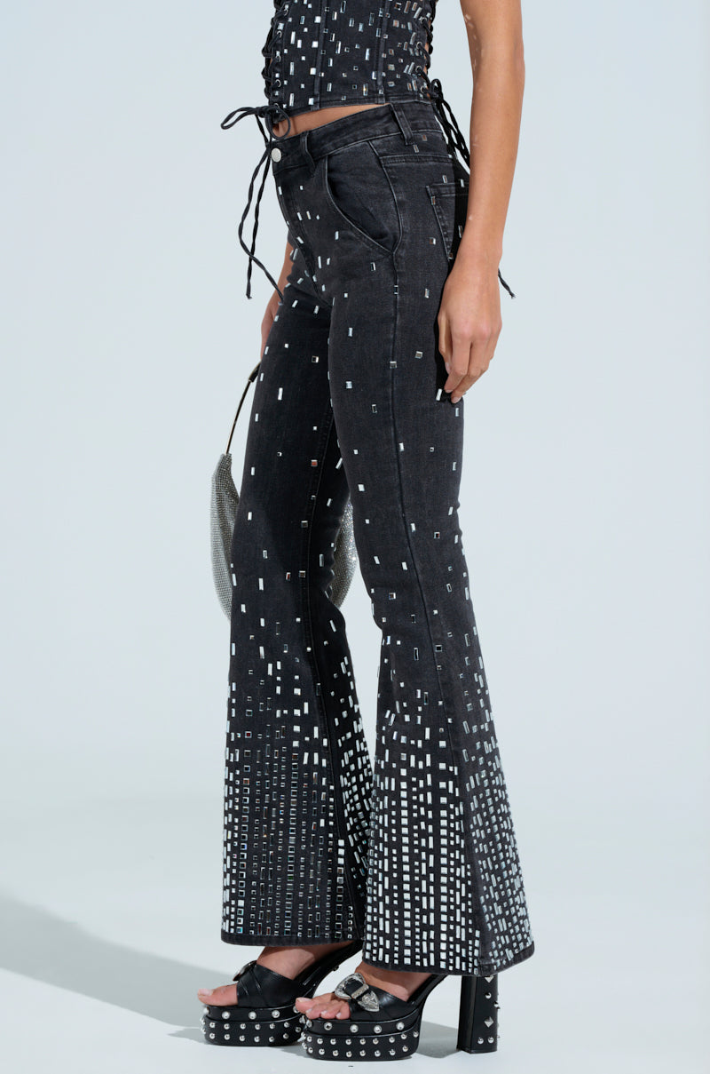 Standing against a plain background, the person showcases black POPSTAR denim pants with a white digital print and a mid-rise waist. They complement it with a matching top, carrying a silver purse and wearing platform sandals embellished with rhinestones for extra flair.