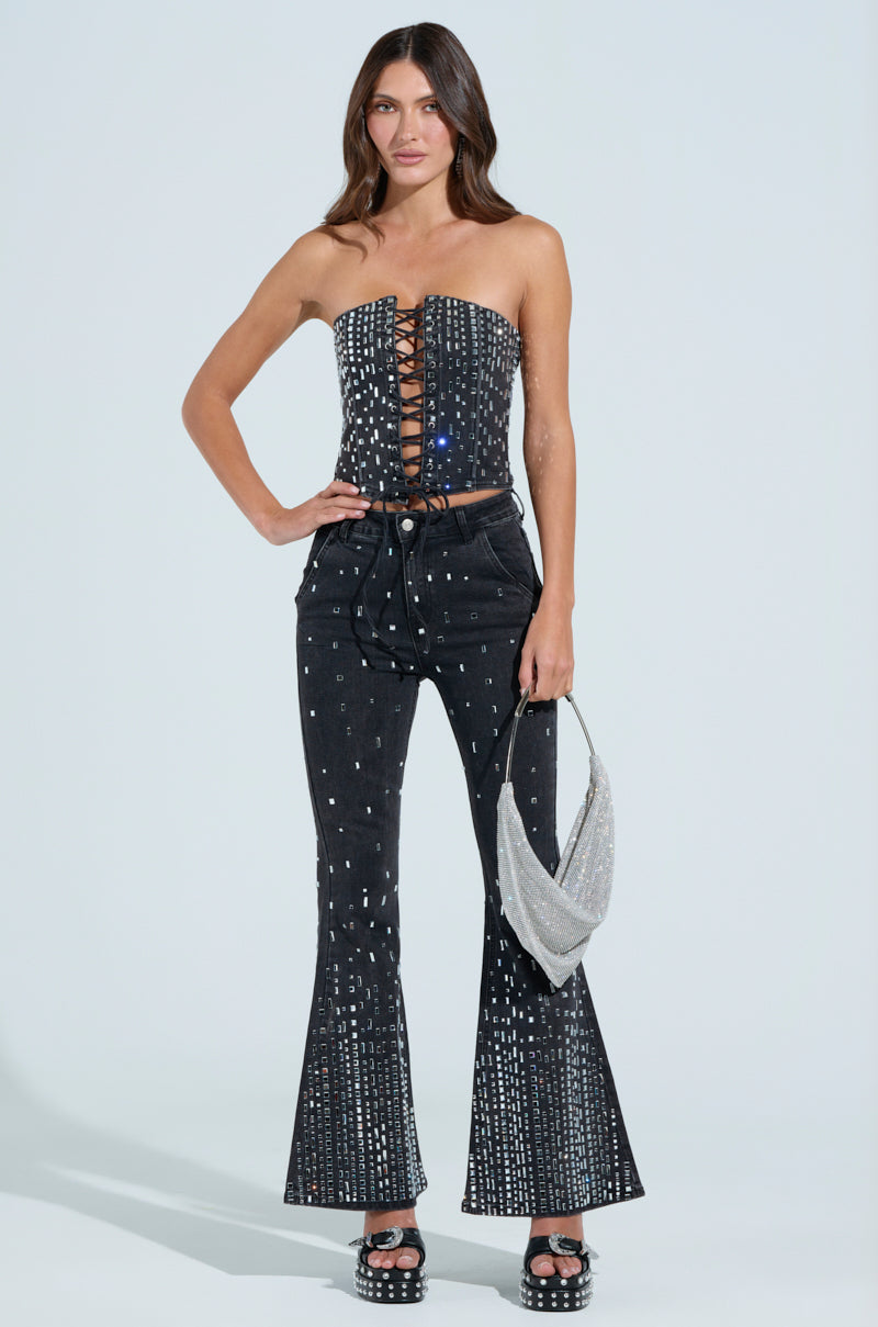 A woman wears a strapless black top with a lace-up front and POPSTAR BLACK DENIM PANT WITH RHINESTONE. She carries a silver purse and dons black platform shoes. Her long, wavy hair perfectly complements her chic look.