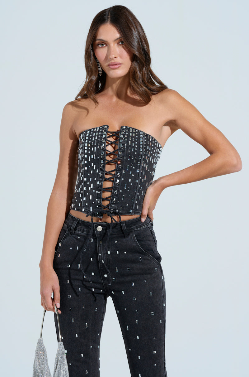 A woman confidently wears the POPSTAR BLACK DENIM CORSET WITH RHINESTONE, featuring a lace-up front and matching pants adorned with rhinestones. She holds a silver purse, and her hair is styled in loose waves.