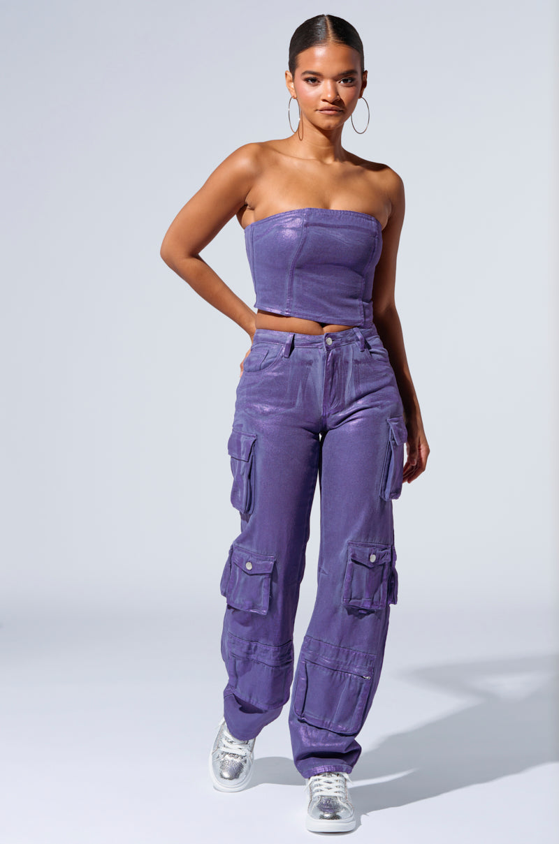 A person with hoop earrings is wearing a strapless purple top and matching THE POWER OF LOVE DENIM CARGO PANT, designed with high-rise, wide-leg features and multiple pockets. They are walking in silver sneakers that add a metallic sheen against the plain, light-colored background. 1