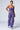 A person with hoop earrings is wearing a strapless purple top and matching THE POWER OF LOVE DENIM CARGO PANT, designed with high-rise, wide-leg features and multiple pockets. They are walking in silver sneakers that add a metallic sheen against the plain, light-colored background.