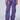 Person wearing THE POWER OF LOVE DENIM CARGO PANT in high-rise wide-leg purple, featuring multiple pockets, paired with metallic silver sneakers, standing against a plain background. Their face is not visible in the image.