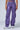 Person wearing THE POWER OF LOVE DENIM CARGO PANT in high-rise wide-leg purple, featuring multiple pockets, paired with metallic silver sneakers, standing against a plain background. Their face is not visible in the image.