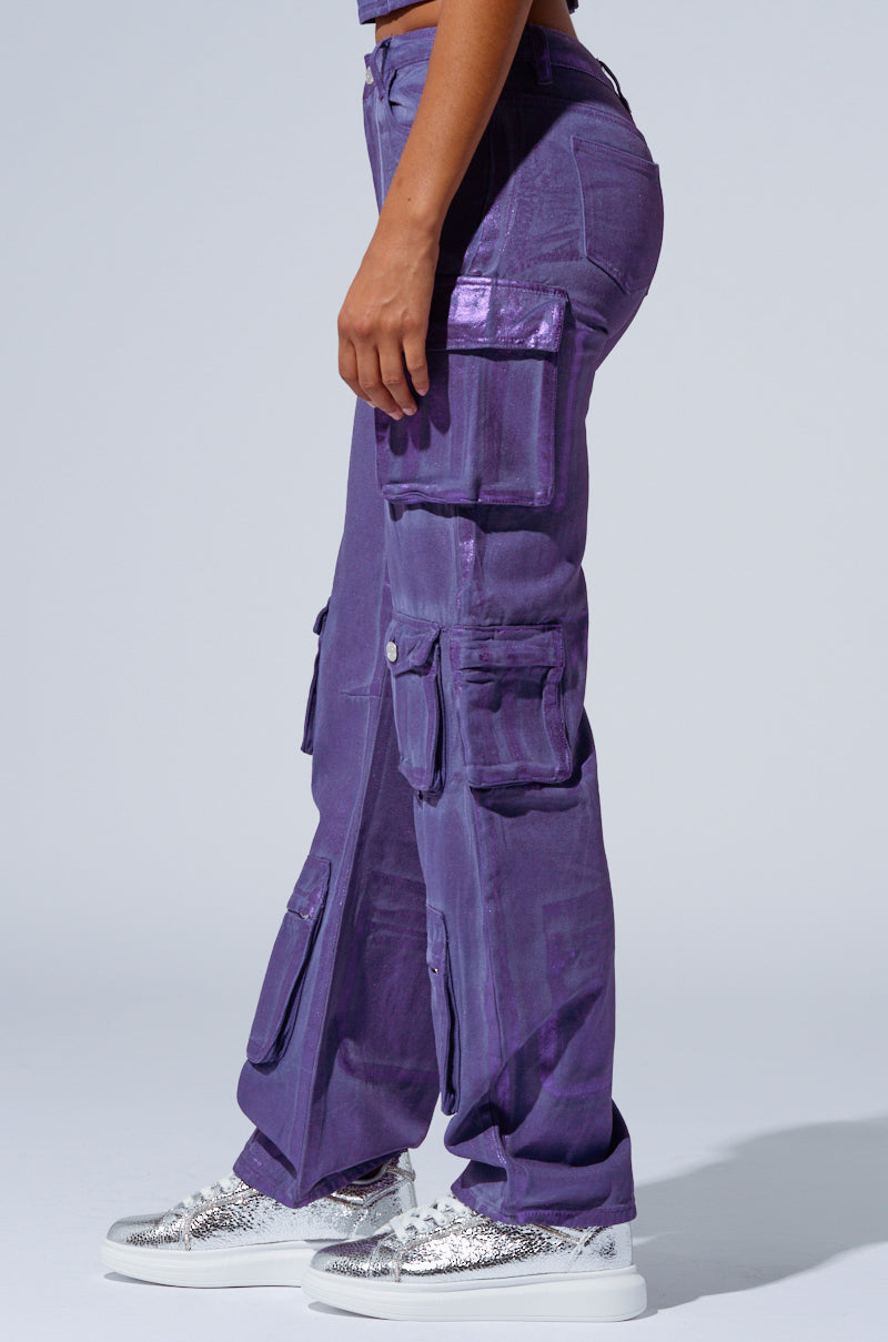 A person is wearing THE POWER OF LOVE DENIM CARGO PANT in a high-rise wide-leg design made of a shiny purple material, featuring multiple pockets. They are also sporting silver glittery sneakers. The background is plain and light-colored. The person is standing sideways, showcasing the right side of their outfit. 2