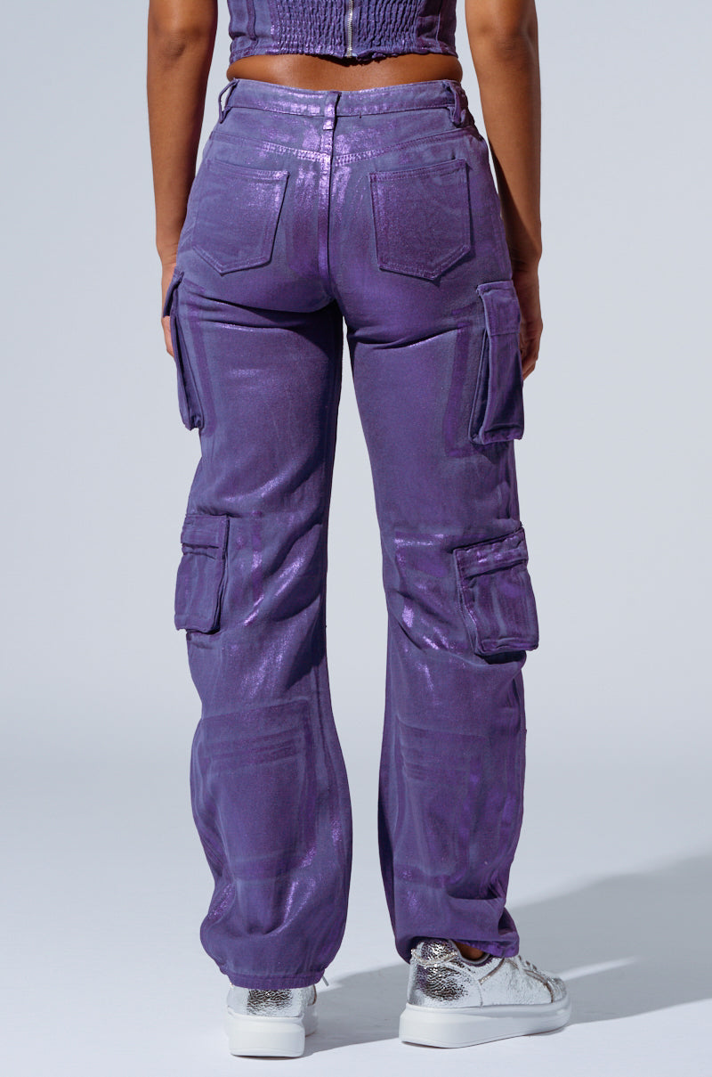A person stands with their back to the camera, donning THE POWER OF LOVE DENIM CARGO PANT in a high-waisted, shiny purple design paired with metallic silver sneakers. The pants feature multiple pockets and detailed stitching. They also sport a matching purple crop top, highlighting a chic outfit with a metallic sheen. 3