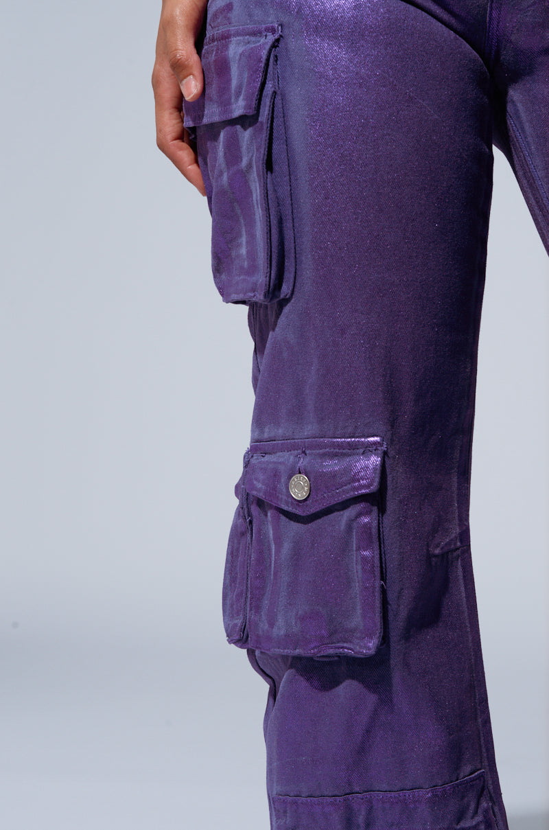 Close-up image of a person wearing THE POWER OF LOVE DENIM CARGO PANT in iridescent purple, featuring multiple pockets. The pants exhibit a slight metallic sheen, with the person’s hand partially visible near the top of the image. The light gray background accentuates the high-rise wide-leg design. 5