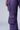 Close-up image of a person wearing THE POWER OF LOVE DENIM CARGO PANT in iridescent purple, featuring multiple pockets. The pants exhibit a slight metallic sheen, with the person’s hand partially visible near the top of the image. The light gray background accentuates the high-rise wide-leg design.