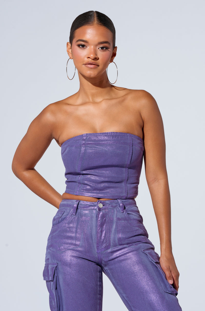 A woman stands confidently against a plain background, wearing THE POWER OF LOVE CORSET TOP, a shiny, purple strapless top with corset-inspired paneling, paired with matching high-waisted pants. She accessorizes with large hoop earrings and has her hair pulled back. One hand rests on her hip.