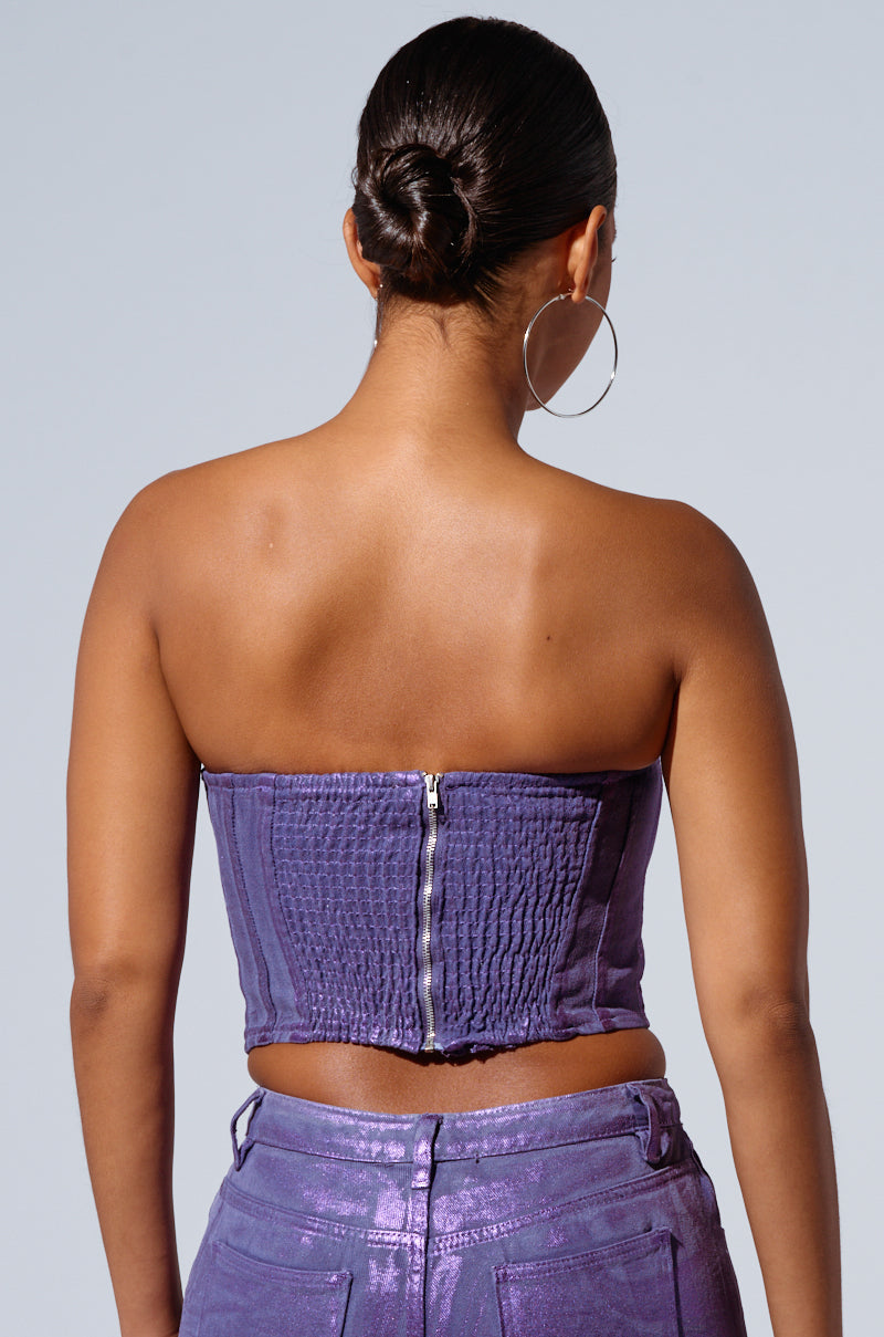 A person is shown from the back, wearing 'THE POWER OF LOVE CORSET TOP' in purple, featuring strapless design and corset-inspired paneling with a central zipper. They are paired with shiny purple pants that have a metallic sheen. Their hair is styled in a sleek, low bun, and they are wearing large hoop earrings. The background is plain and light gray.
