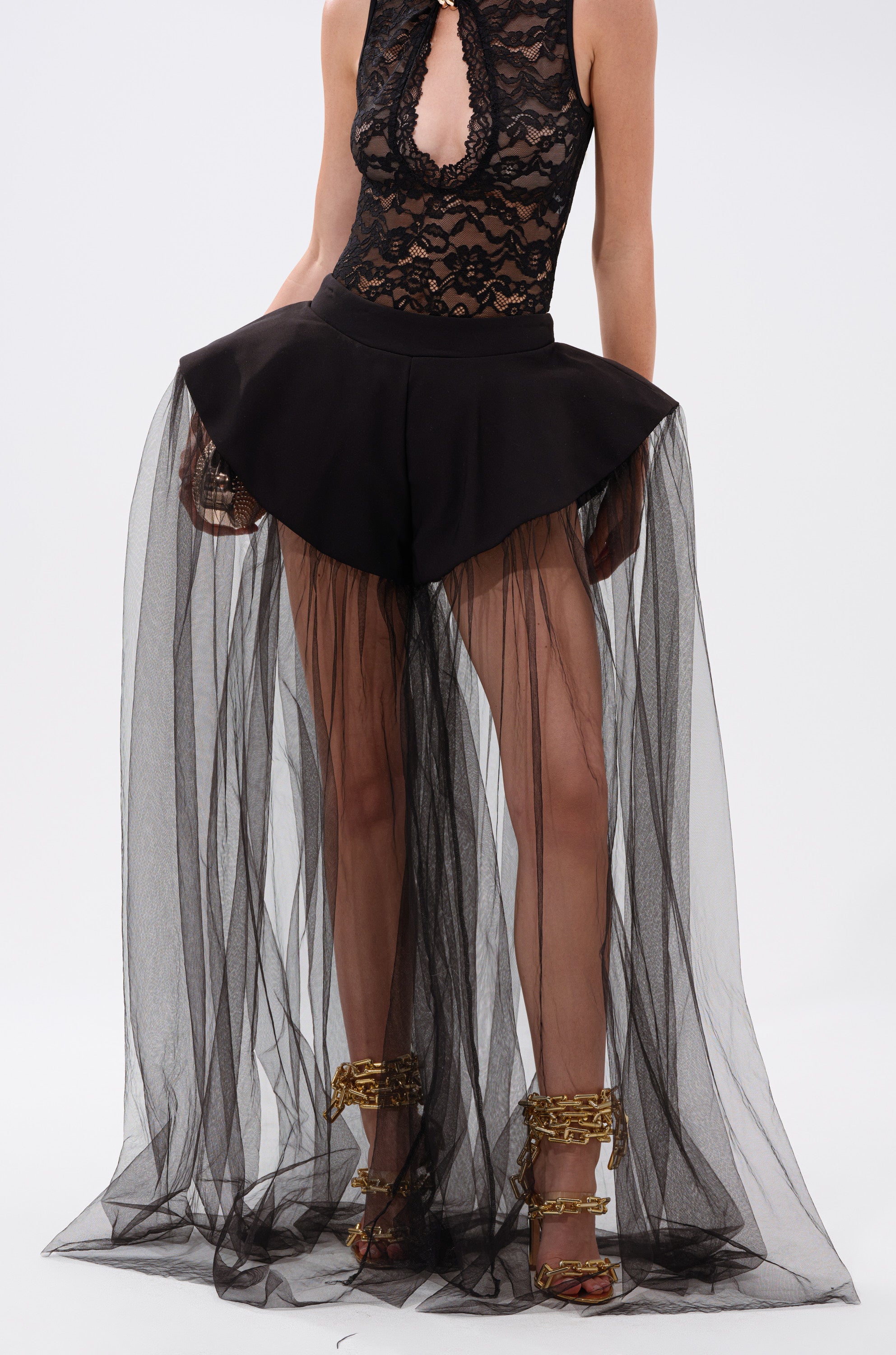 Wearing the ALL OF THE DRAMA TULLE PANTS, styled with a black lace sleeveless bodysuit and gold chain-embellished heels, featuring high-waist shorts and a sheer black tulle avant-garde overskirt.