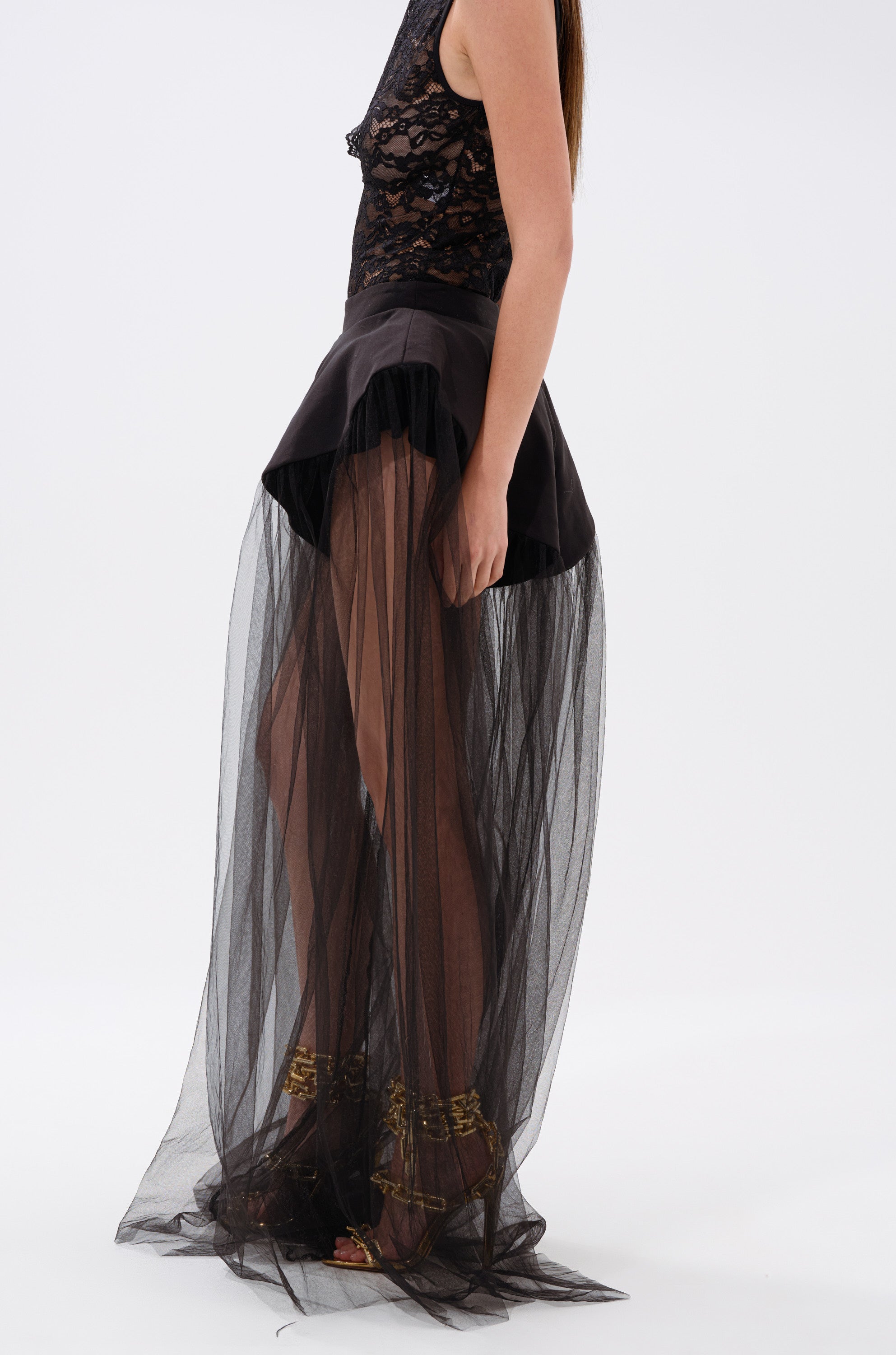 A person showcases avant-garde style in the ALL OF THE DRAMA TULLE PANTS—sheer black tulle layered over a short skirt—paired with gold high-heeled sandals, set against a plain white background.