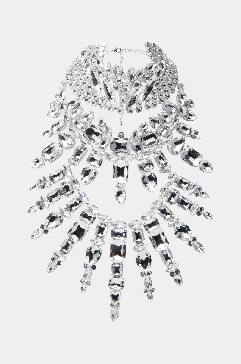 IM THE STATEMENT NECKLACE boasts a triple-layer design with large clear crystal gems and rhinestone accents in intricate patterns, delivering an ornate, dramatic look against a light gray background.