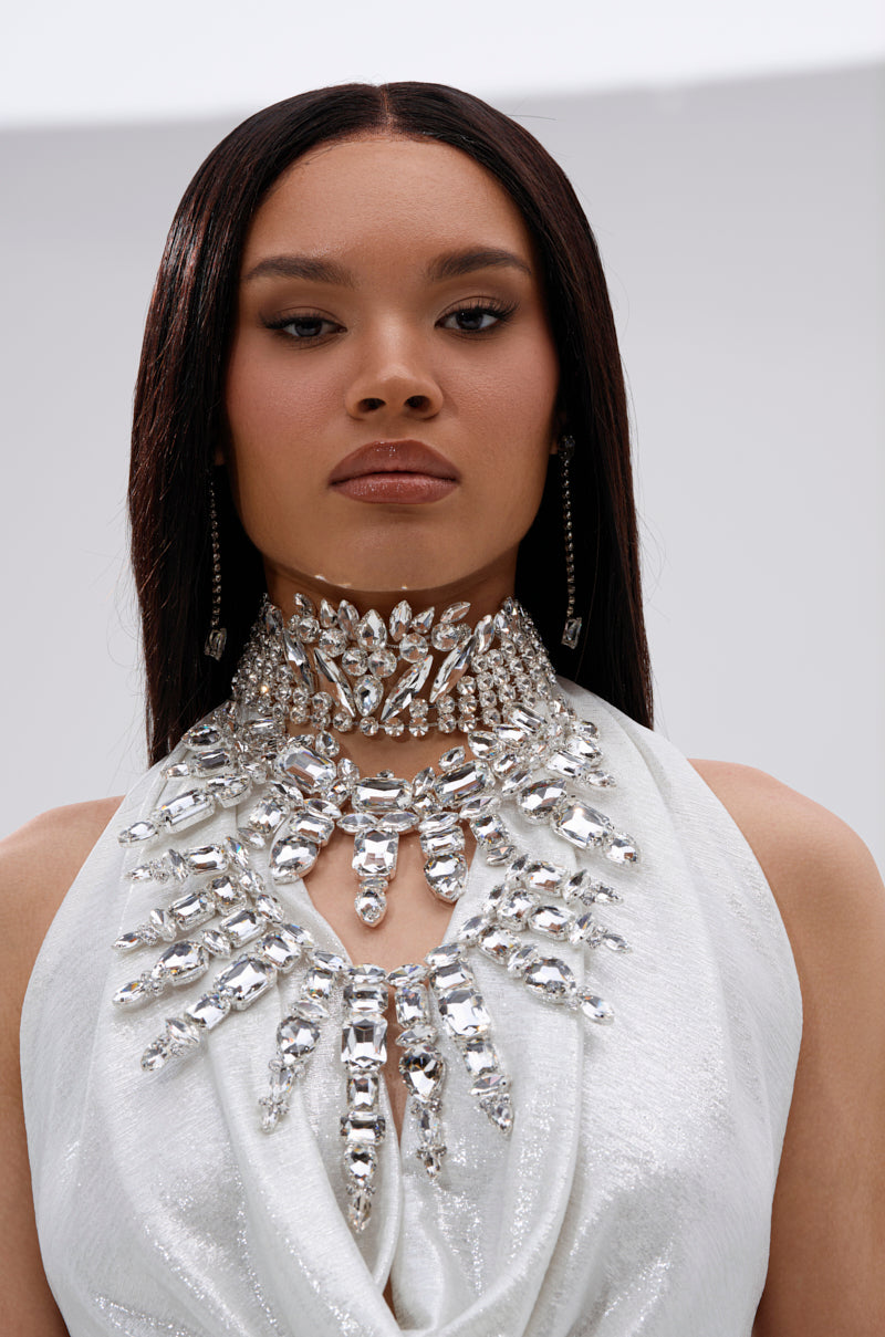 A woman with straight dark hair wears a shimmering white outfit and the IM THE STATEMENT NECKLACE, adorned with rhinestones. She gazes directly at the camera, her expression neutral, against a simple backdrop.