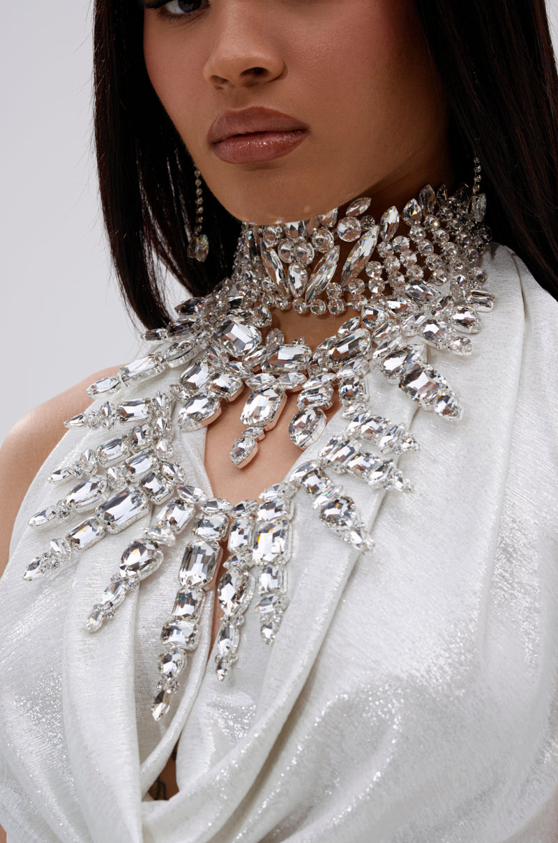 A woman wears the IM THE STATEMENT NECKLACE with matching rhinestone earrings, both sparkling in the light. She pairs them with a shimmering white halter top, while the image focuses on her lower face, neck, and upper chest.