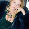 A woman with long blonde hair is wearing a dark blue fur coat and a green sequined top, accessorized with the BOOK THE TRIP STATEMENT NECKLACE, featuring faux gold accents. The background is white adorned with black floral designs, as she gazes directly at the camera.