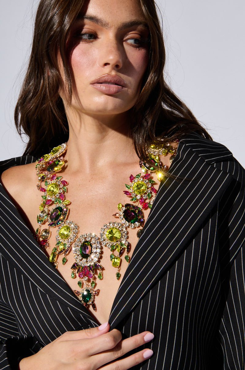 A woman with long brown hair and fair skin is wearing the BOOK THE TRIP STATEMENT NECKLACE, adorned with colorful gemstones and rhinestone detailing. She is dressed in a black pinstriped blazer, slightly open at the front to showcase the stunning piece against a plain white background. 1