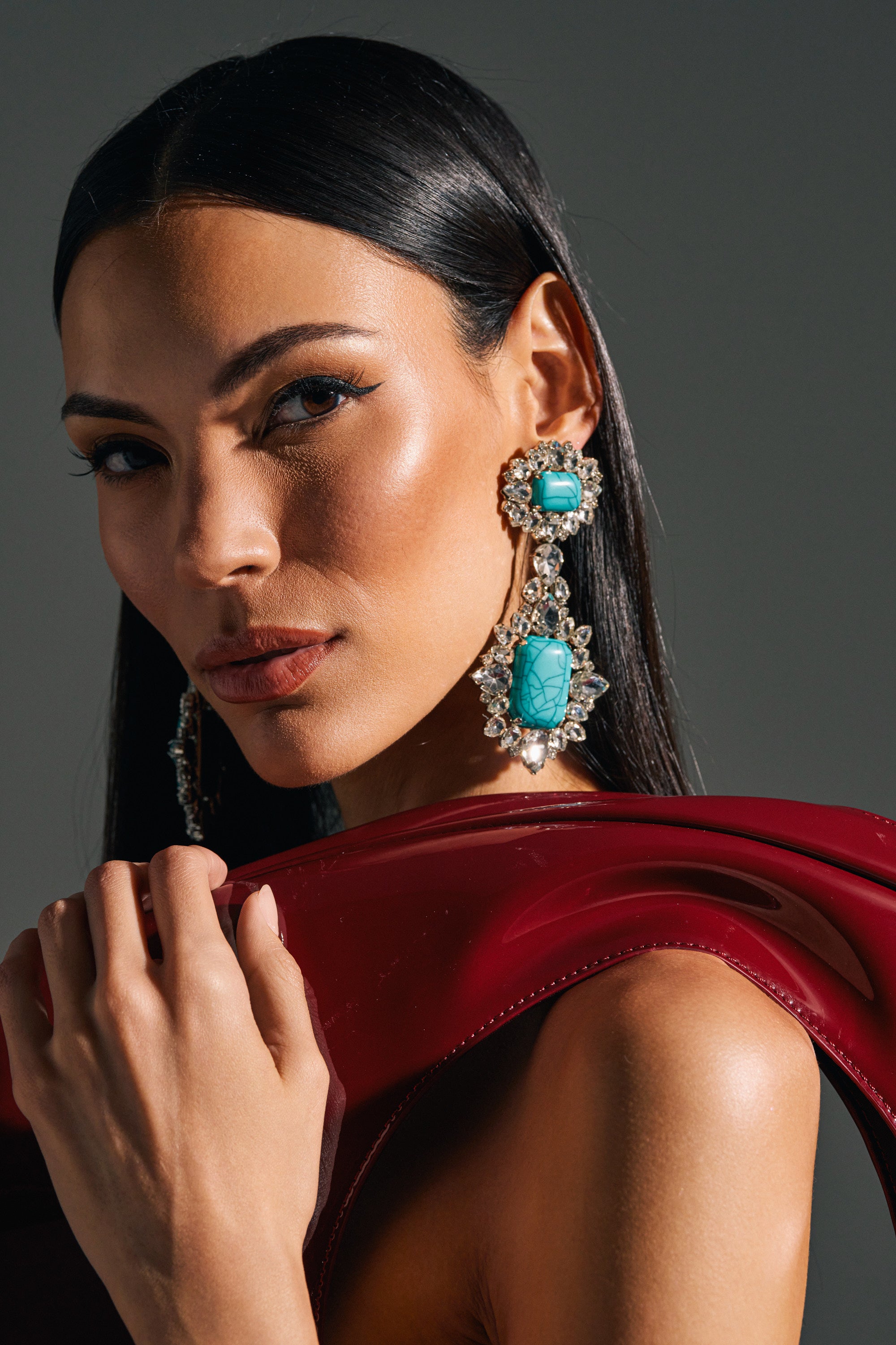 A woman with sleek black hair wears TOTALLY TURQ EARRING statement dangles in turquoise and crystal, holding a shiny red garment over her shoulder as she confidently gazes at the camera against a plain background.