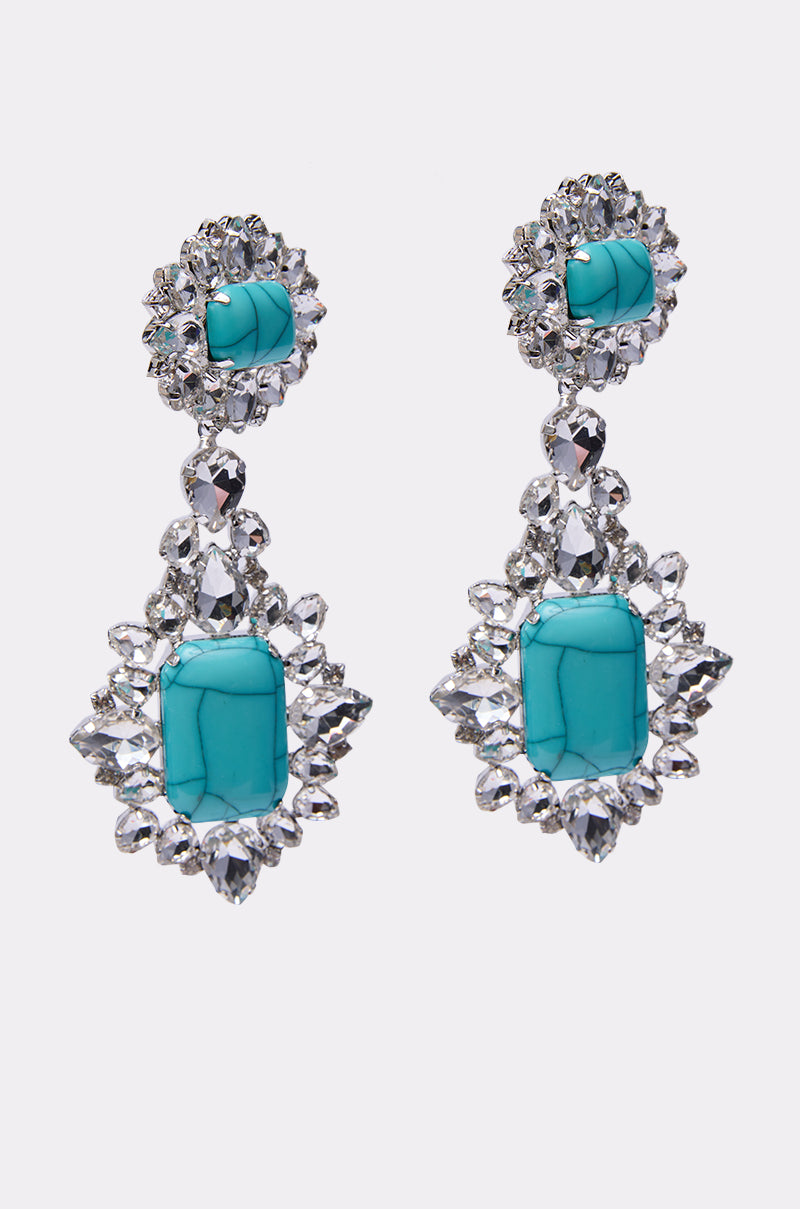 The TOTALLY TURQ EARRING is a pair of ornate statement dangle earrings with turquoise center stones, surrounded by sparkling clear crystals and rhinestone accents in an intricate symmetrical design on a white background.