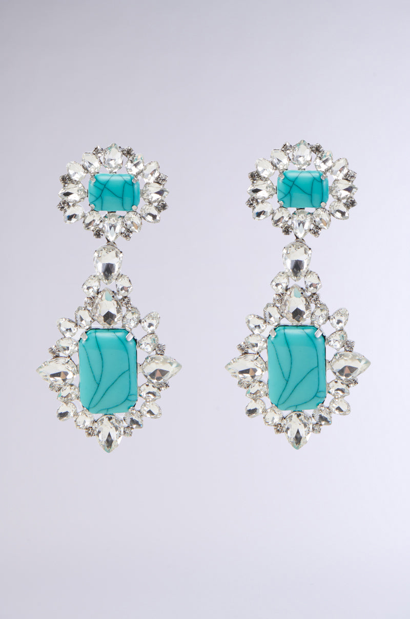 Introducing the TOTALLY TURQ EARRING by GARY WOOD: a pair of elegant, silver-toned drop earrings featuring marbled turquoise gemstones surrounded by sparkling, clear rhinestones. The design includes a smaller square gem at the top and a larger rectangular gem below, both adorned with intricate rhinestone settings.