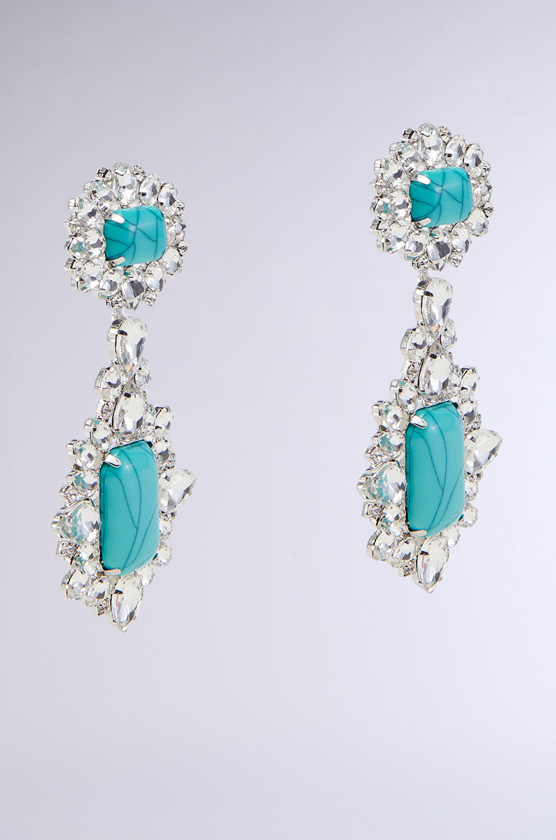 Introducing the TOTALLY TURQ EARRING by GARY WOOD: an exquisite pair of ornate drop earrings adorned with vibrant turquoise gemstones and clusters of sparkling clear crystals. These earrings feature rhinestone embellishments and a marbled gemstone design, showcasing a floral motif with smaller stones at the top and larger ones dangling below, all set against a light grey background.